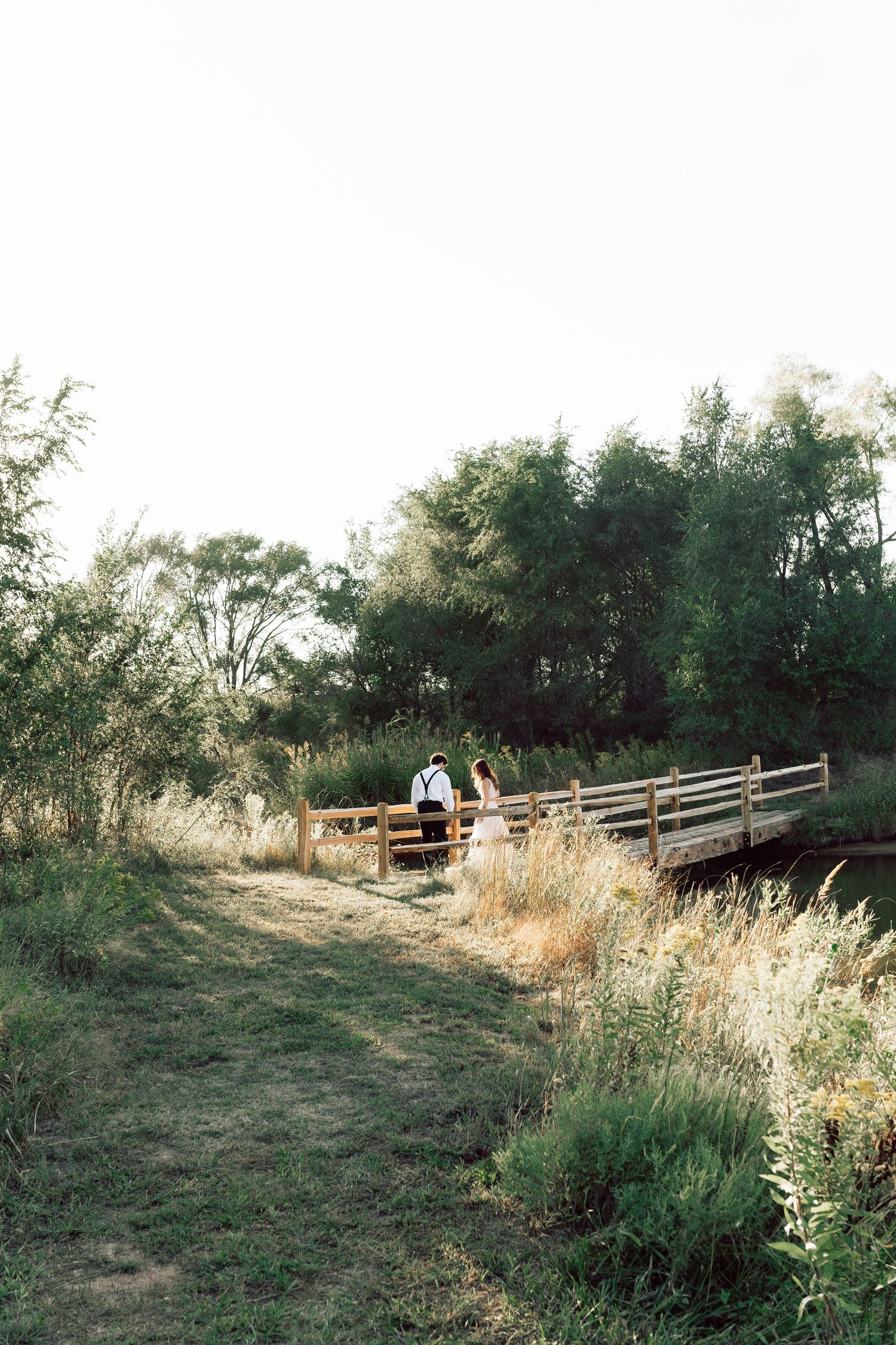 Rustic wooden bridge spans across calm water surrounded by lush greenery at sunset.