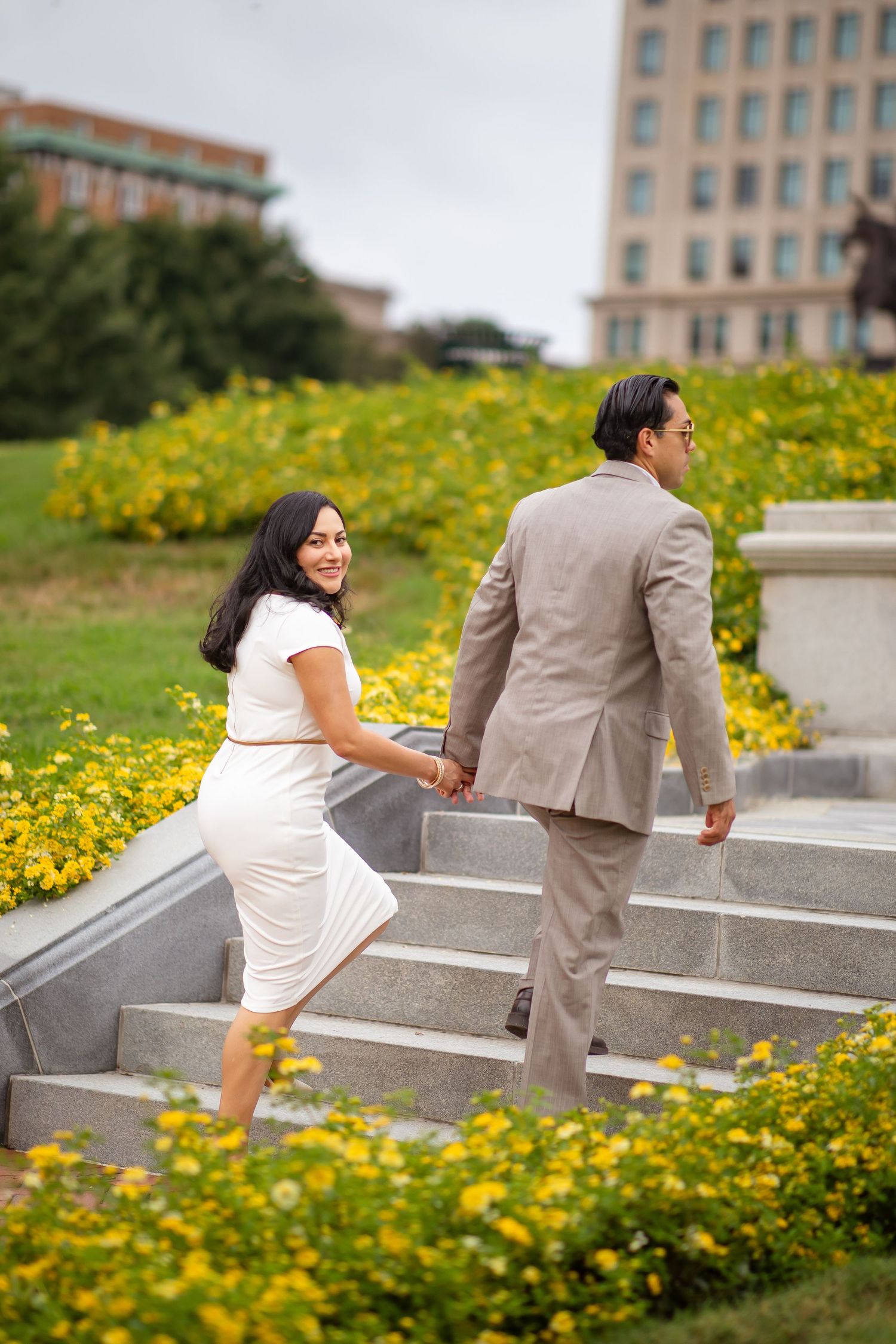 Couple holding hands on stone steps surrounded by yellow flowers, woman in white dress smiling back at camera.