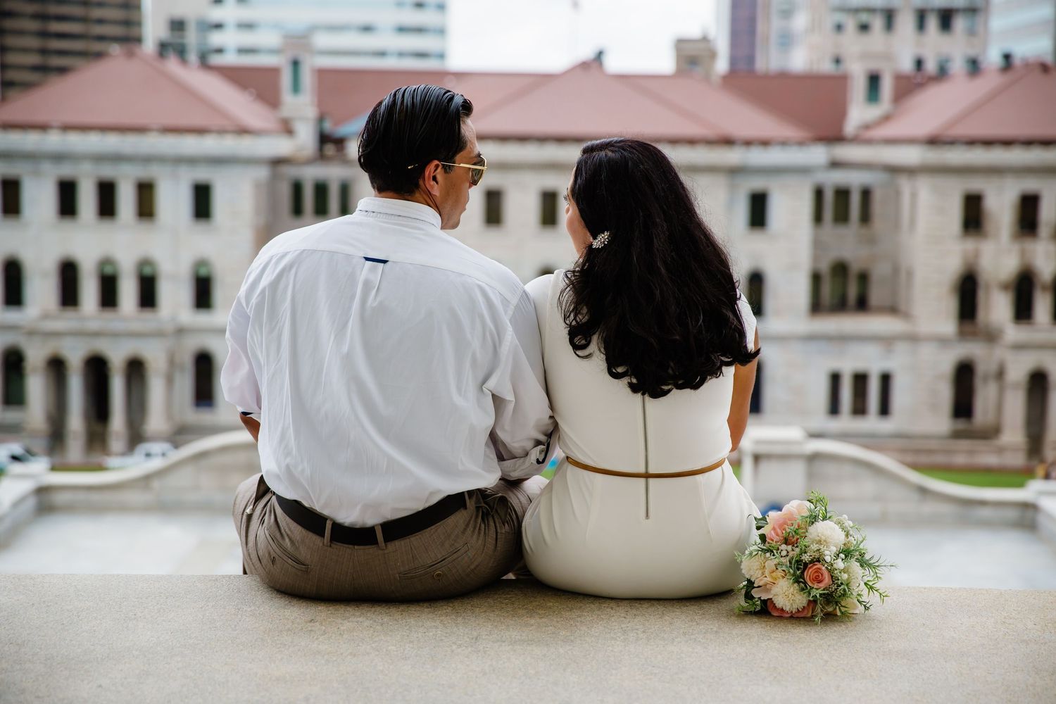 Couple sitting together on a ledge with a bridal bouquet, viewed from behind, with a classical building in the background.