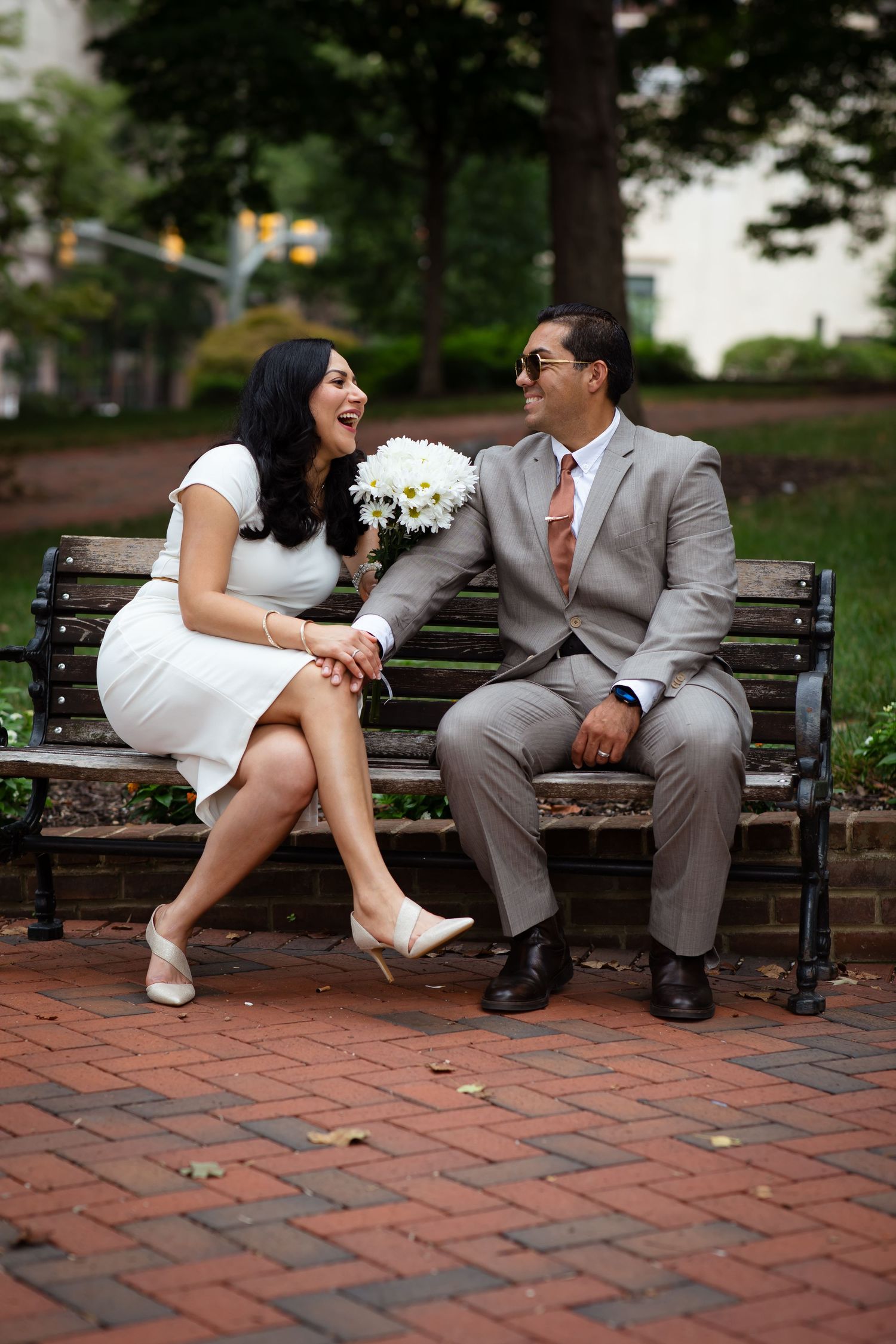 A laughing couple in wedding attire sits on a park bench, woman in white dress holding daisy bouquet, man in gray suit.