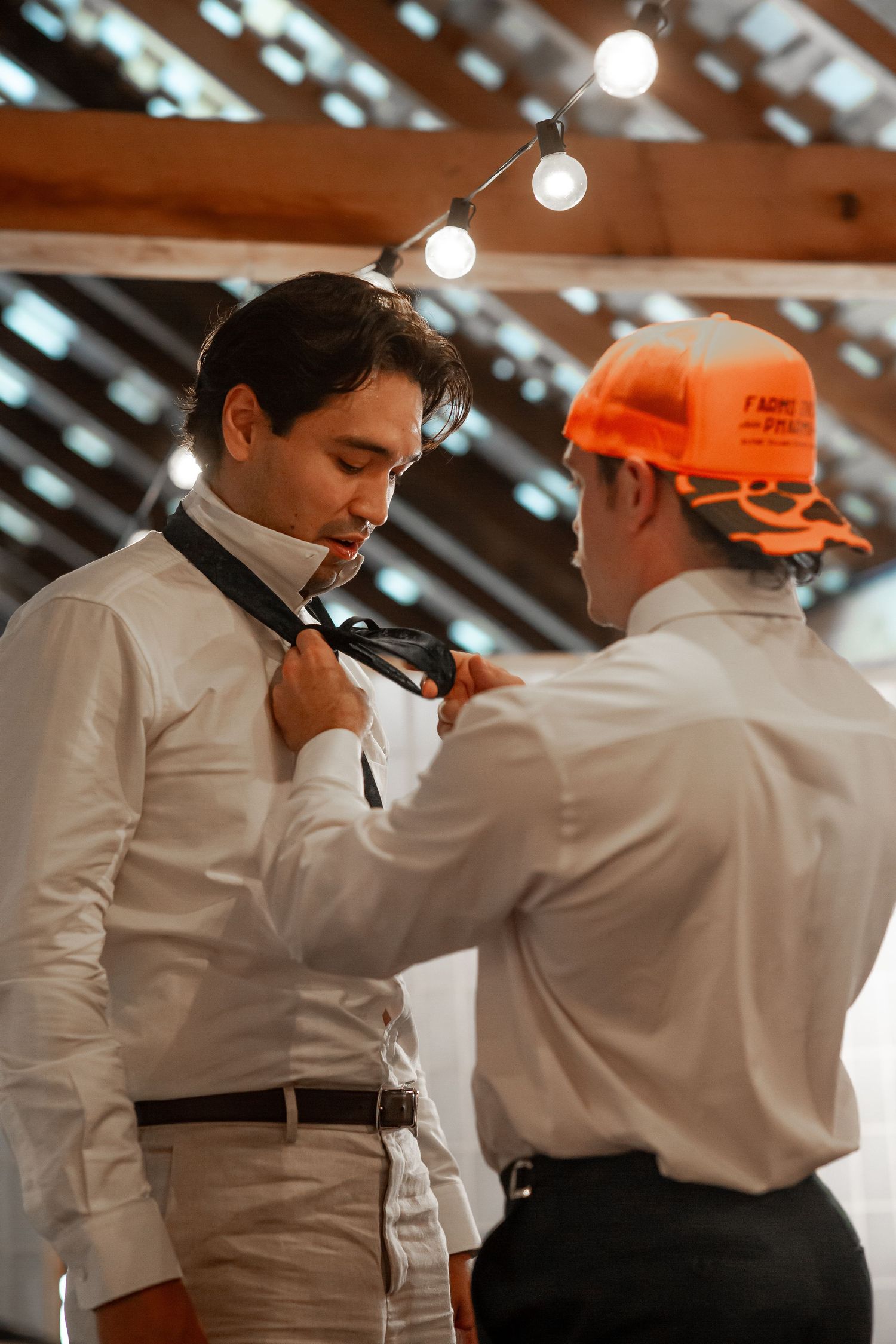 A man in an orange cap helps adjust another man's tie while both wear white dress shirts indoors.