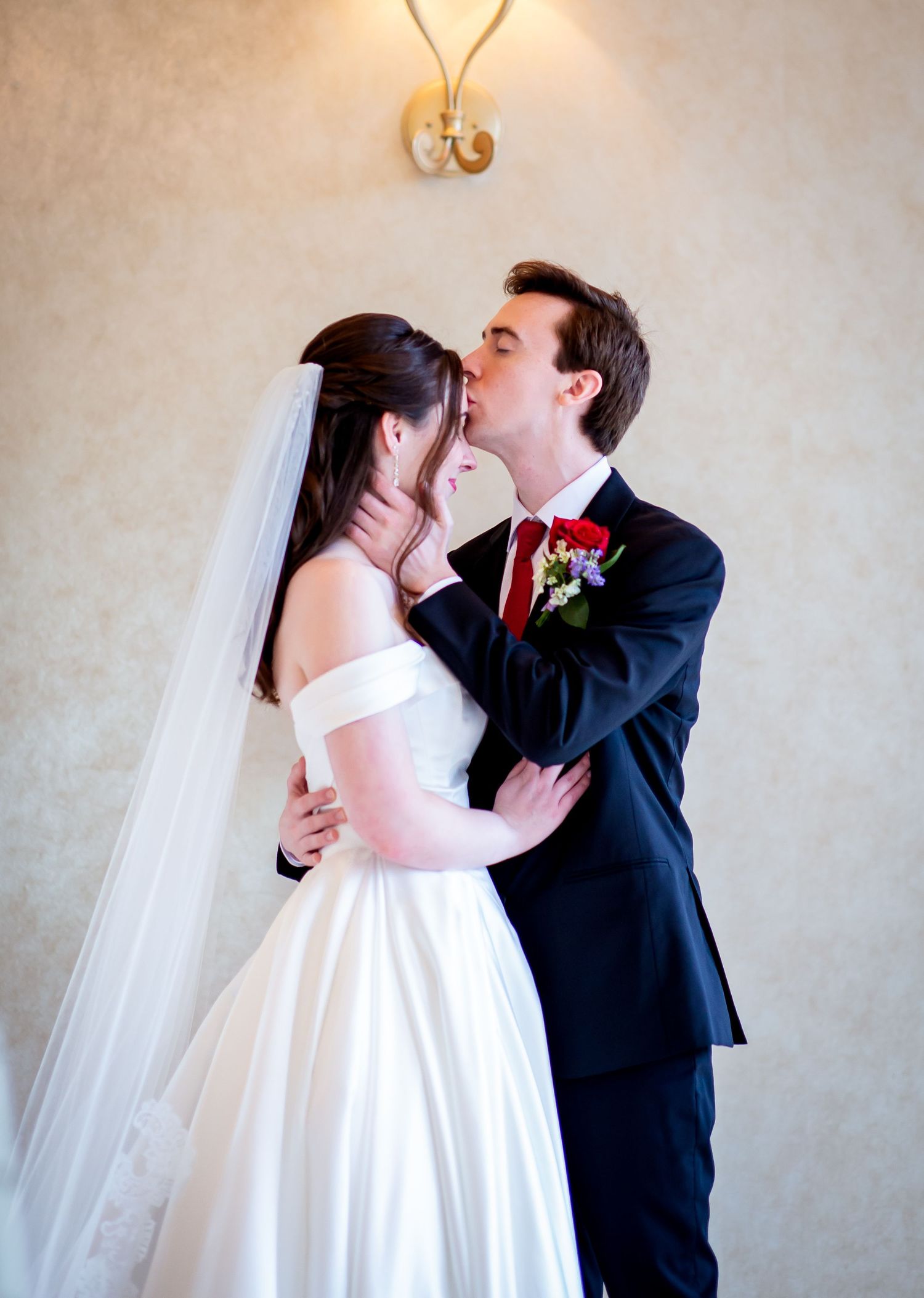 Groom in navy suit kisses bride in off-shoulder white gown and veil during romantic wedding portrait.