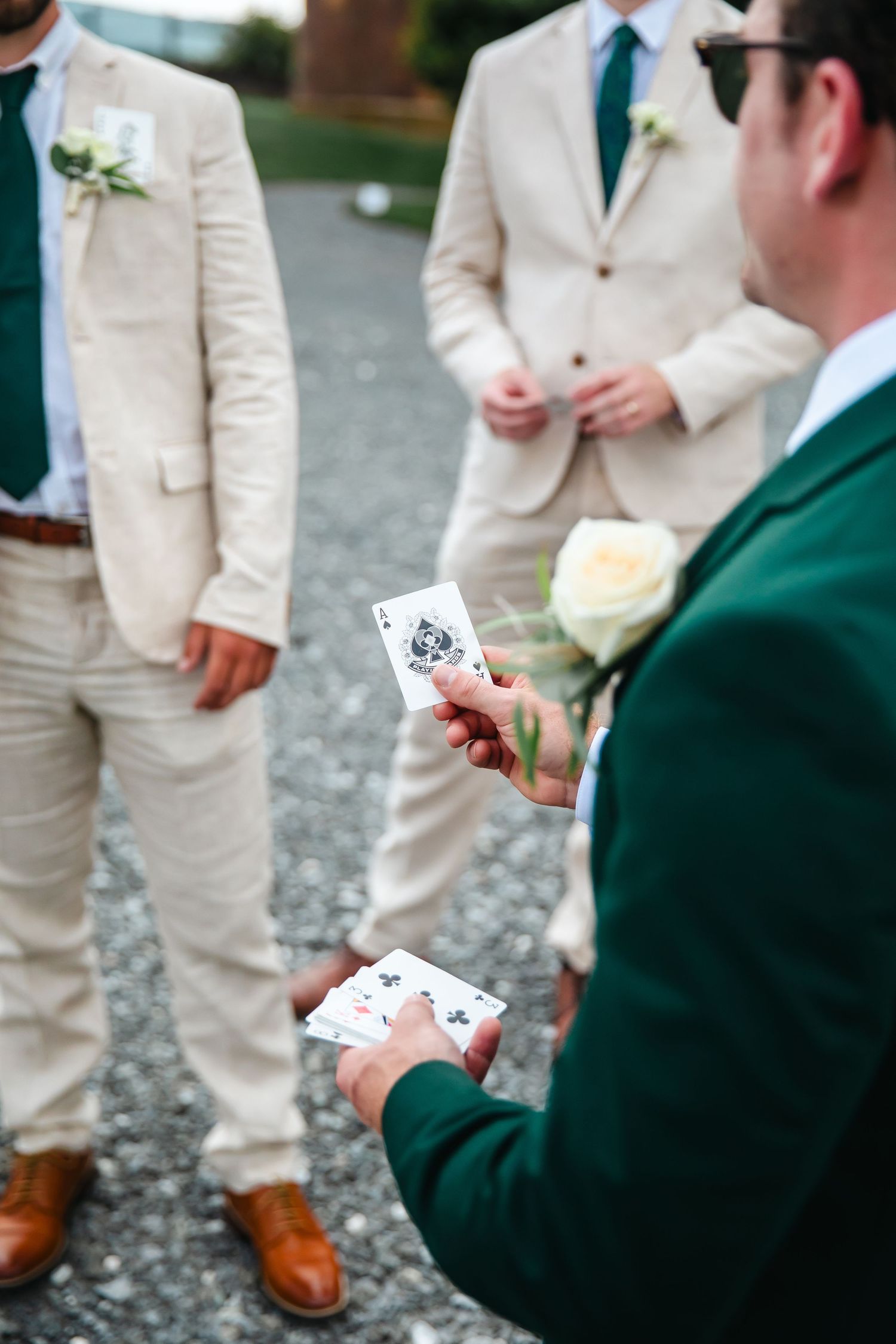 Groomsmen in tan suits and green jackets playing cards outdoors at a wedding, holding playing cards with boutonnieres.