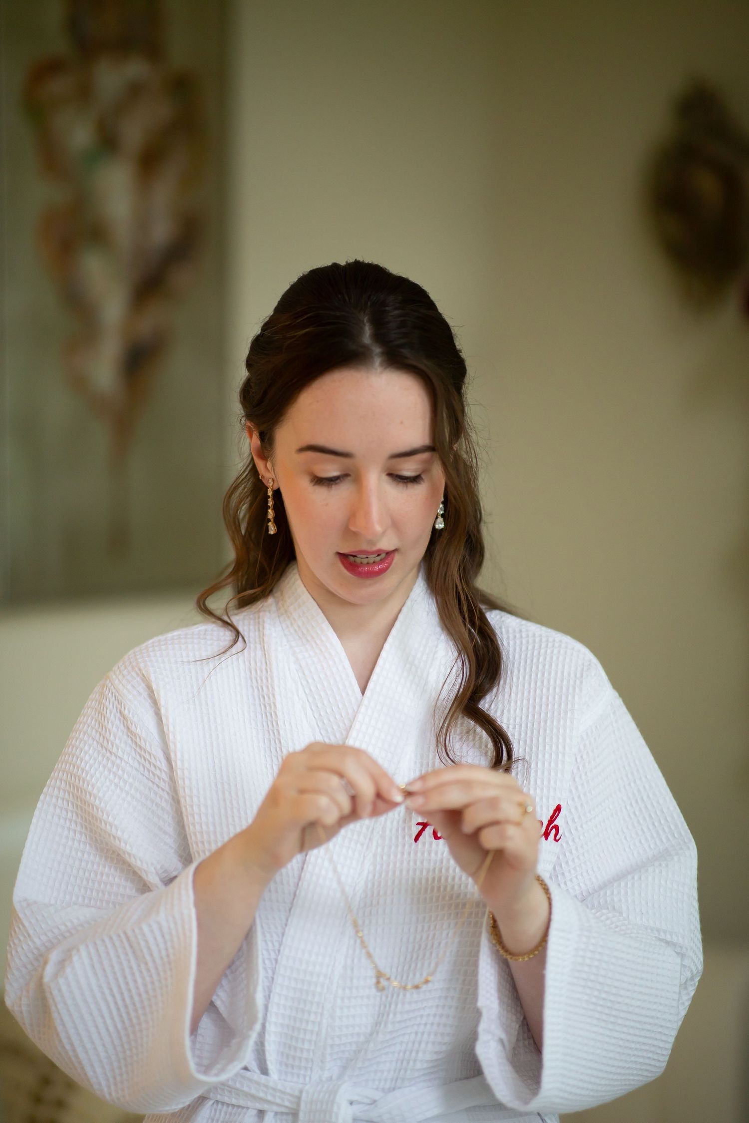 Beautiful bride in white monogrammed robe fastening delicate necklace before wedding.