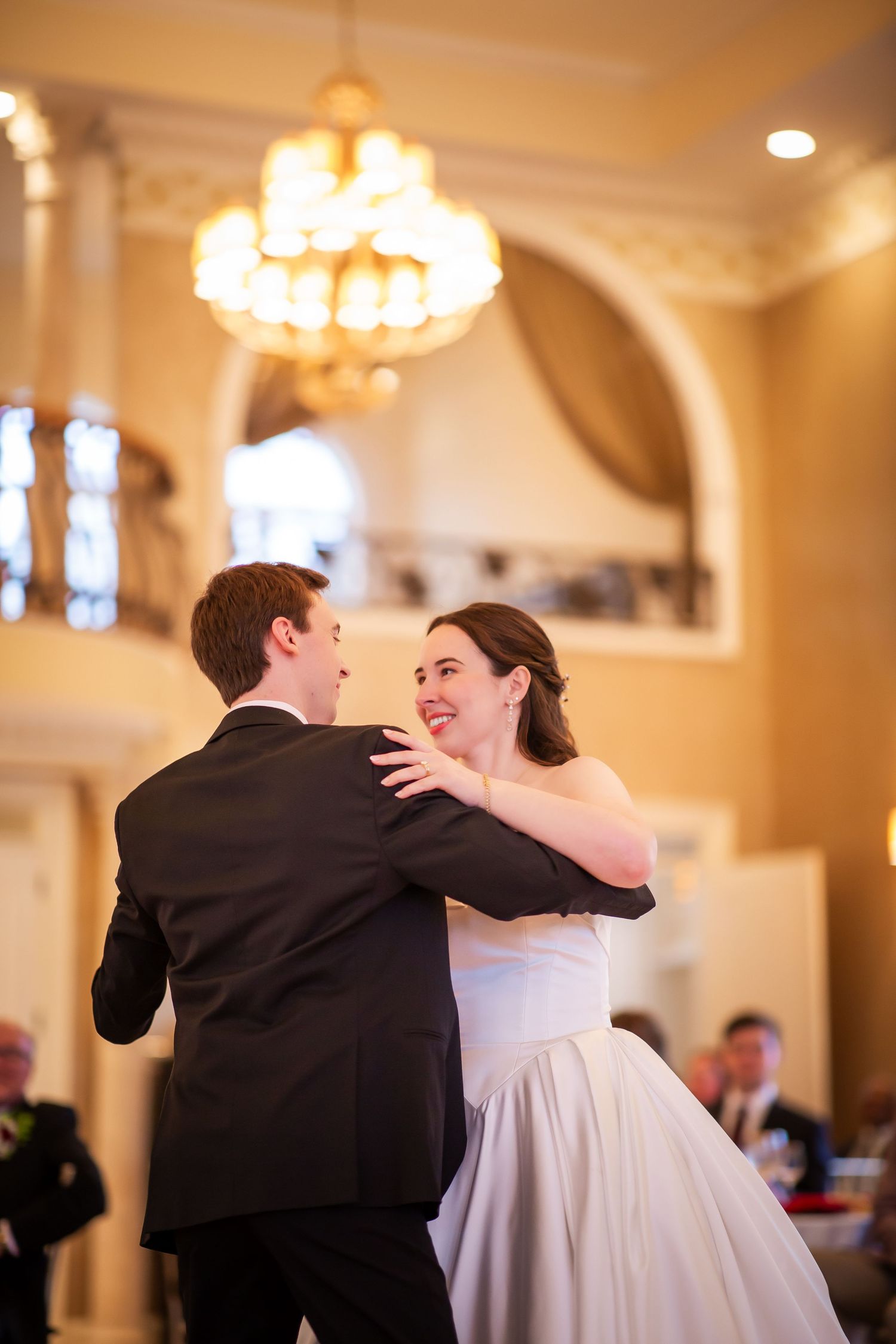 Bride laughs joyfully while sharing first dance with groom beneath golden chandelier at wedding reception.