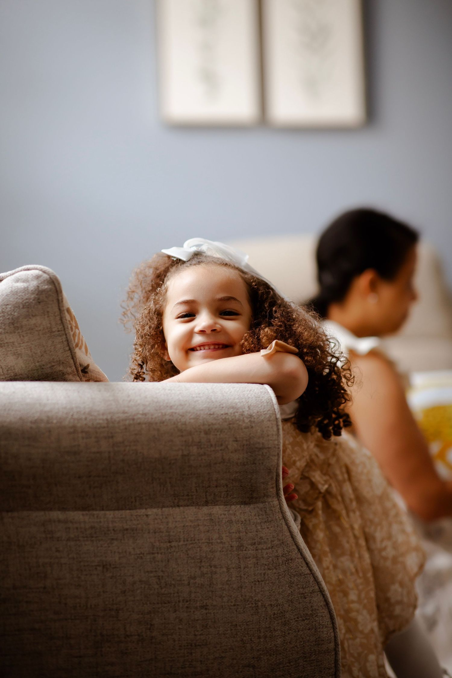 Little girl leans on couch and smiles at camera with mom in the background