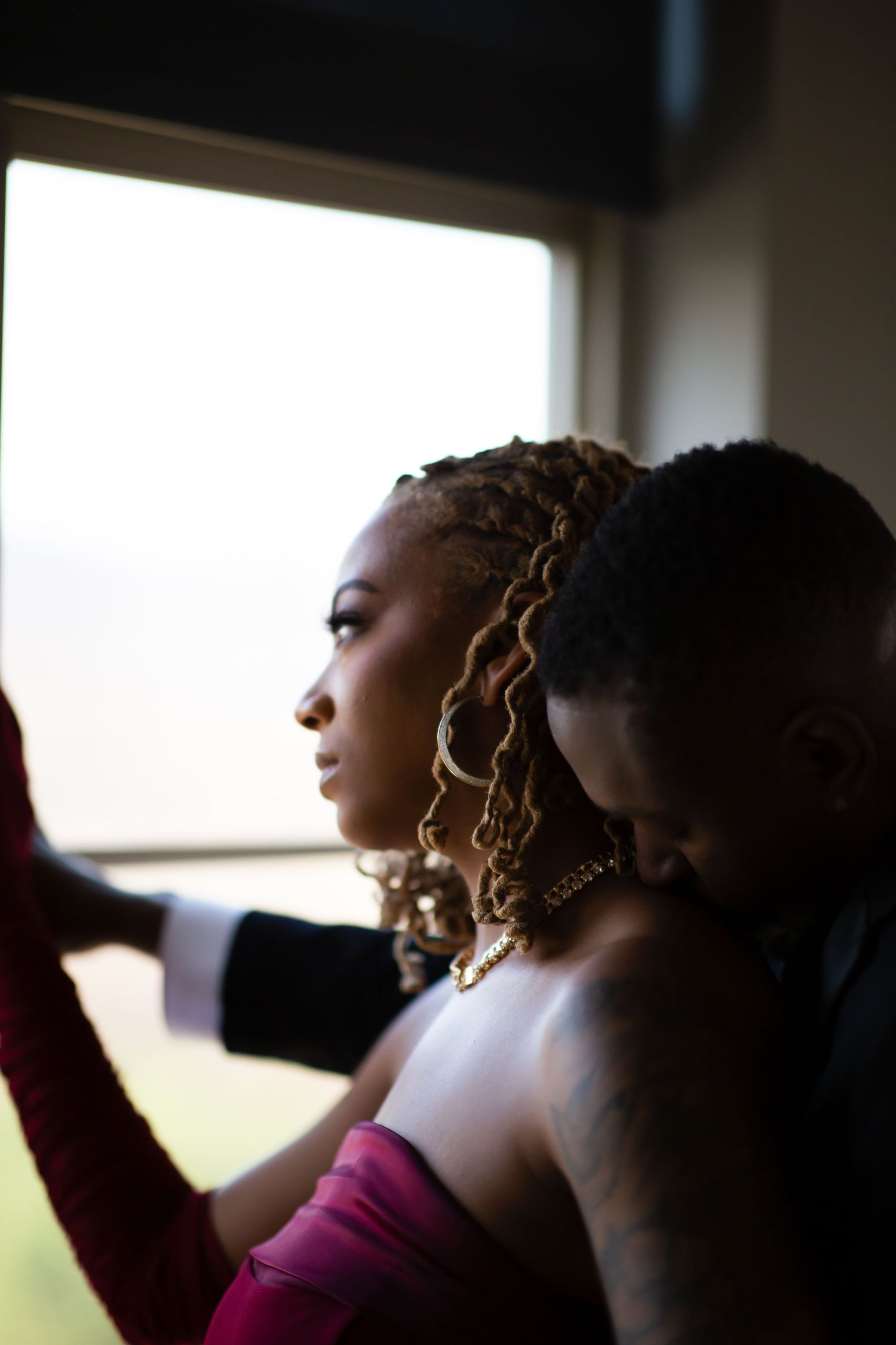 A Black couple gazes out a window together, the woman in a red strapless gown with locs.