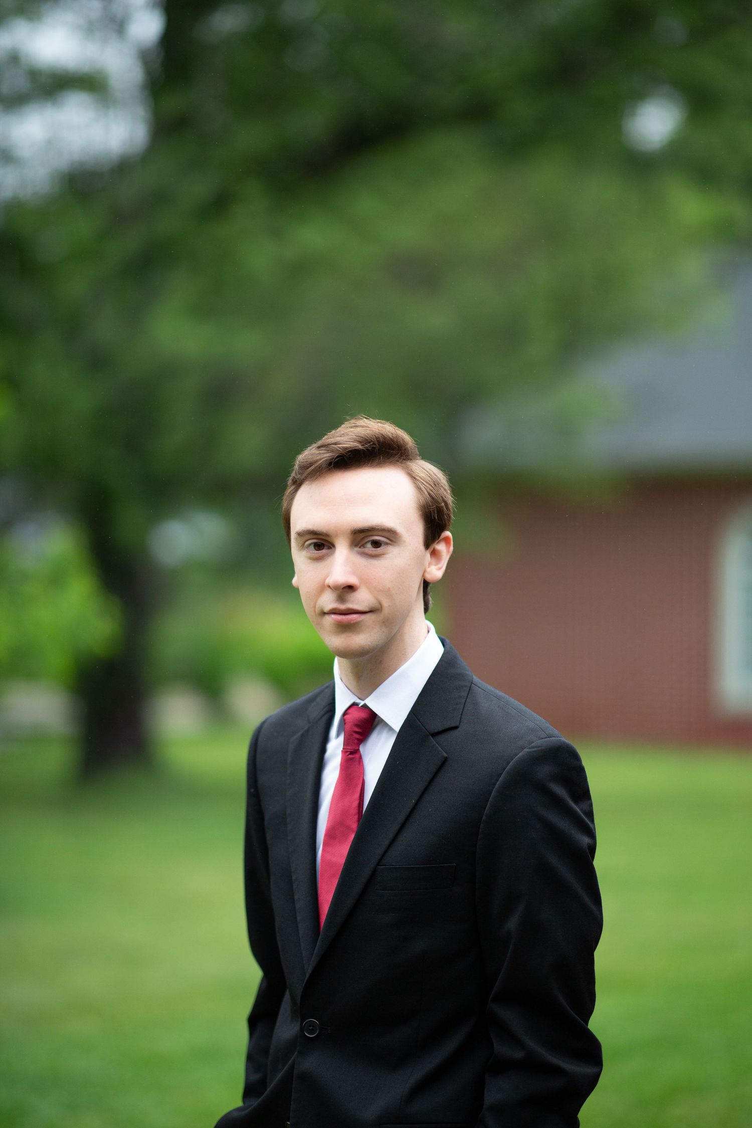 Young groom in black suit and red tie posing outdoors with green trees and brick building background.