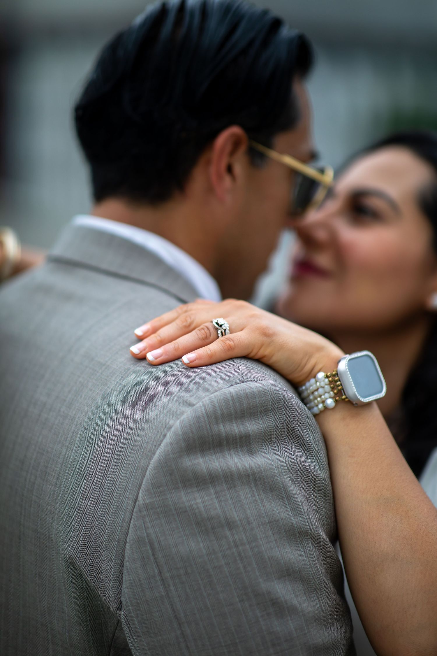 Couple in elegant attire embracing closely, woman wearing a pearl bracelet, smartwatch, and diamond ring on her hand.