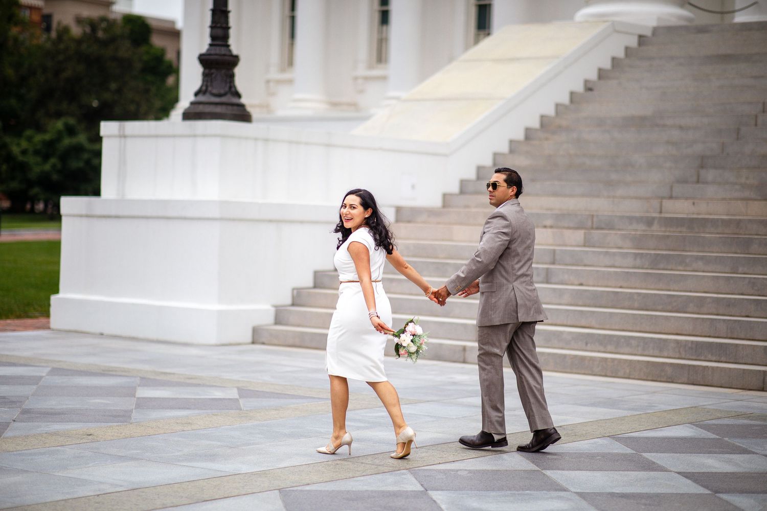 Bride in white dress and groom in gray suit hold hands walking near white marble steps at outdoor wedding venue.
