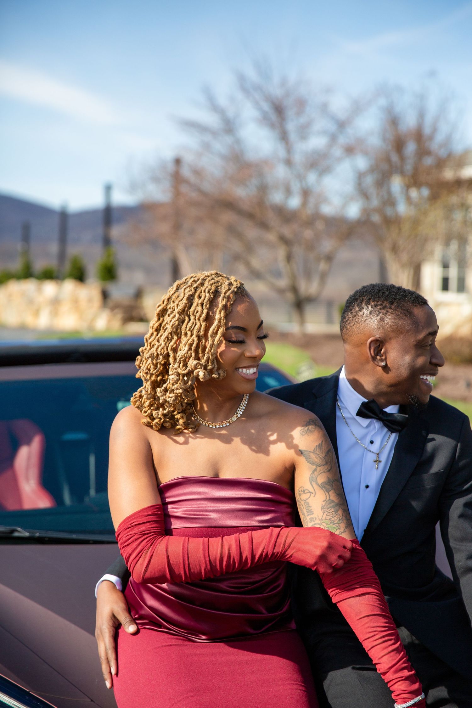 Stylish couple leaning against a red convertible, woman in strapless burgundy dress with red gloves, man in black tuxedo.