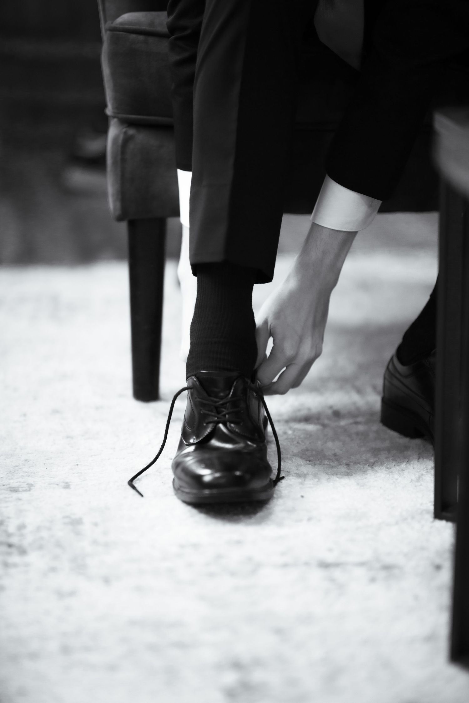 Groom tying black dress shoe laces while sitting in chair before wedding ceremony.