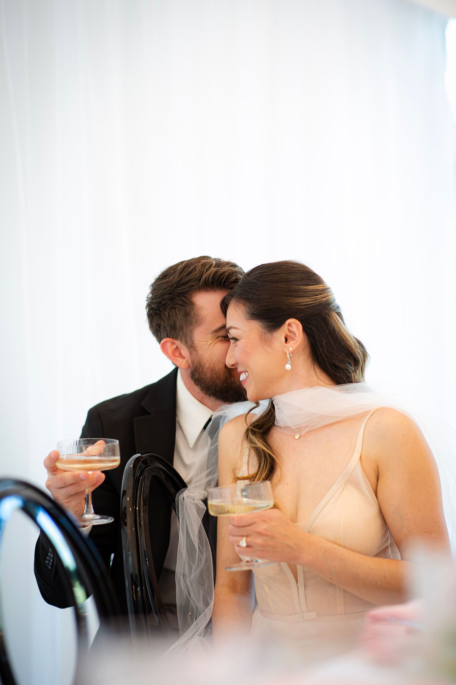 Bride leaning in to groom as he whispers in her ear while drinking champagne