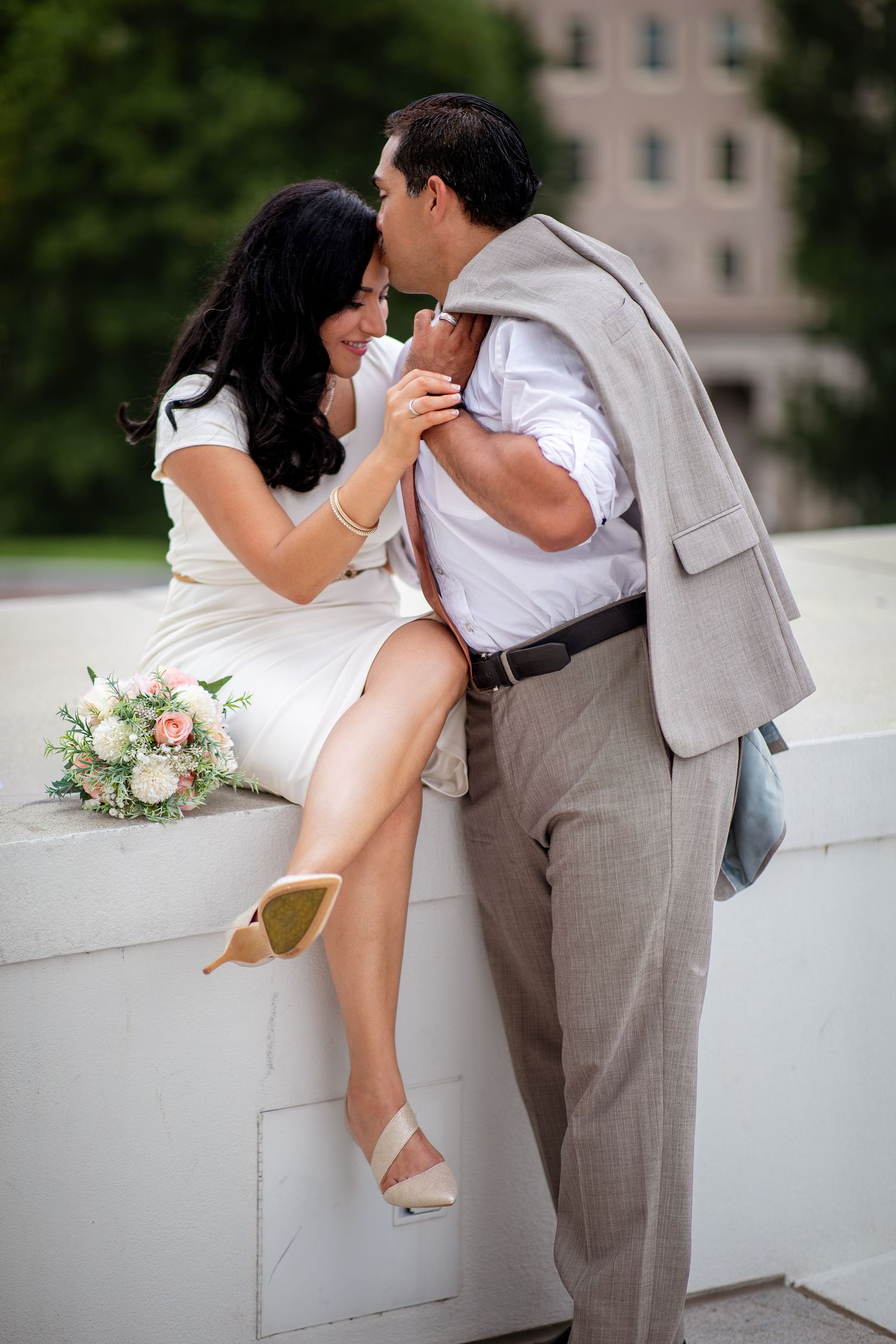 Romantic couple sharing a tender kiss at their wedding, bride in white dress holding a bouquet, groom in gray suit.