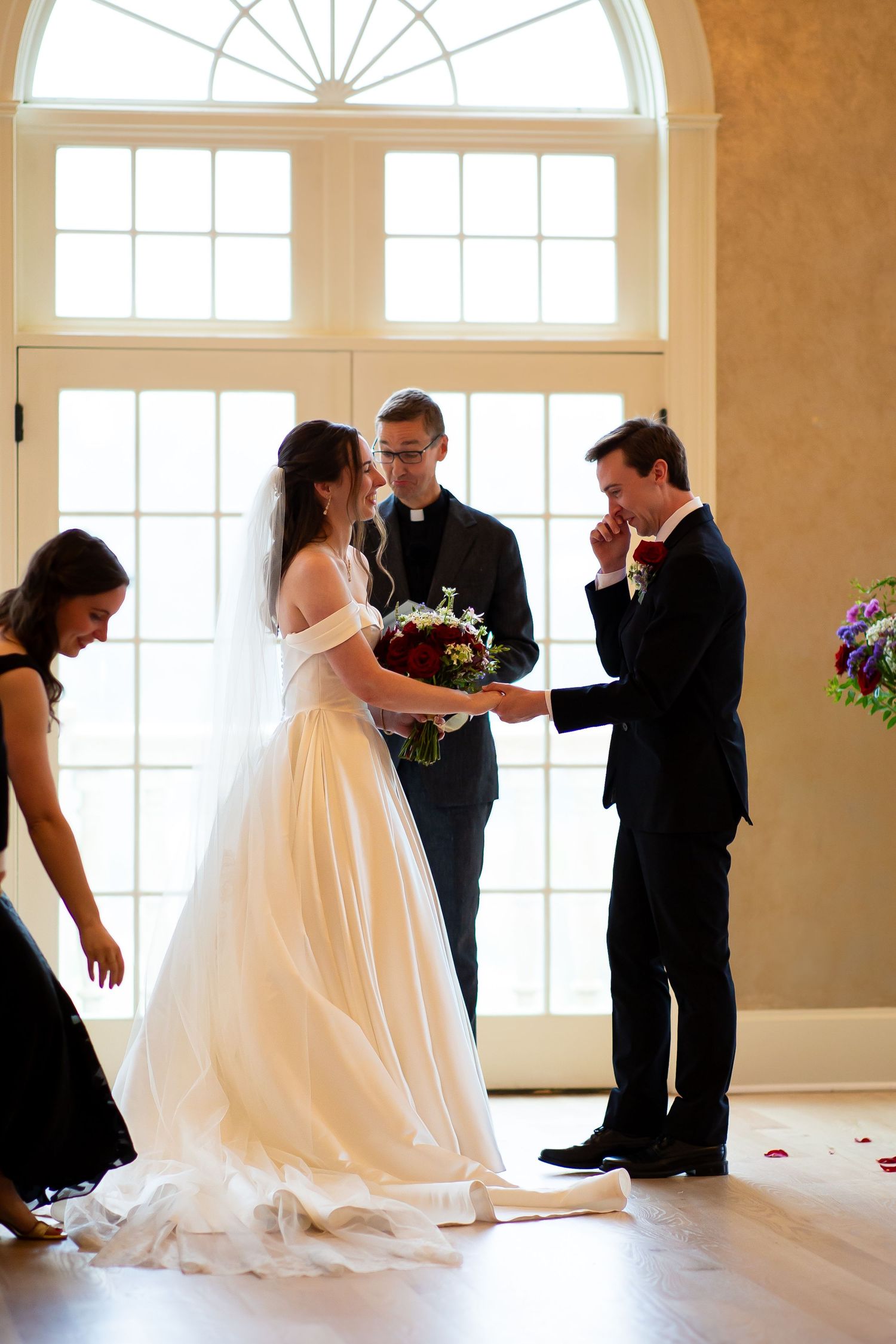 An emotional groom wipes tears as the bride holds his hands during their indoor wedding ceremony.