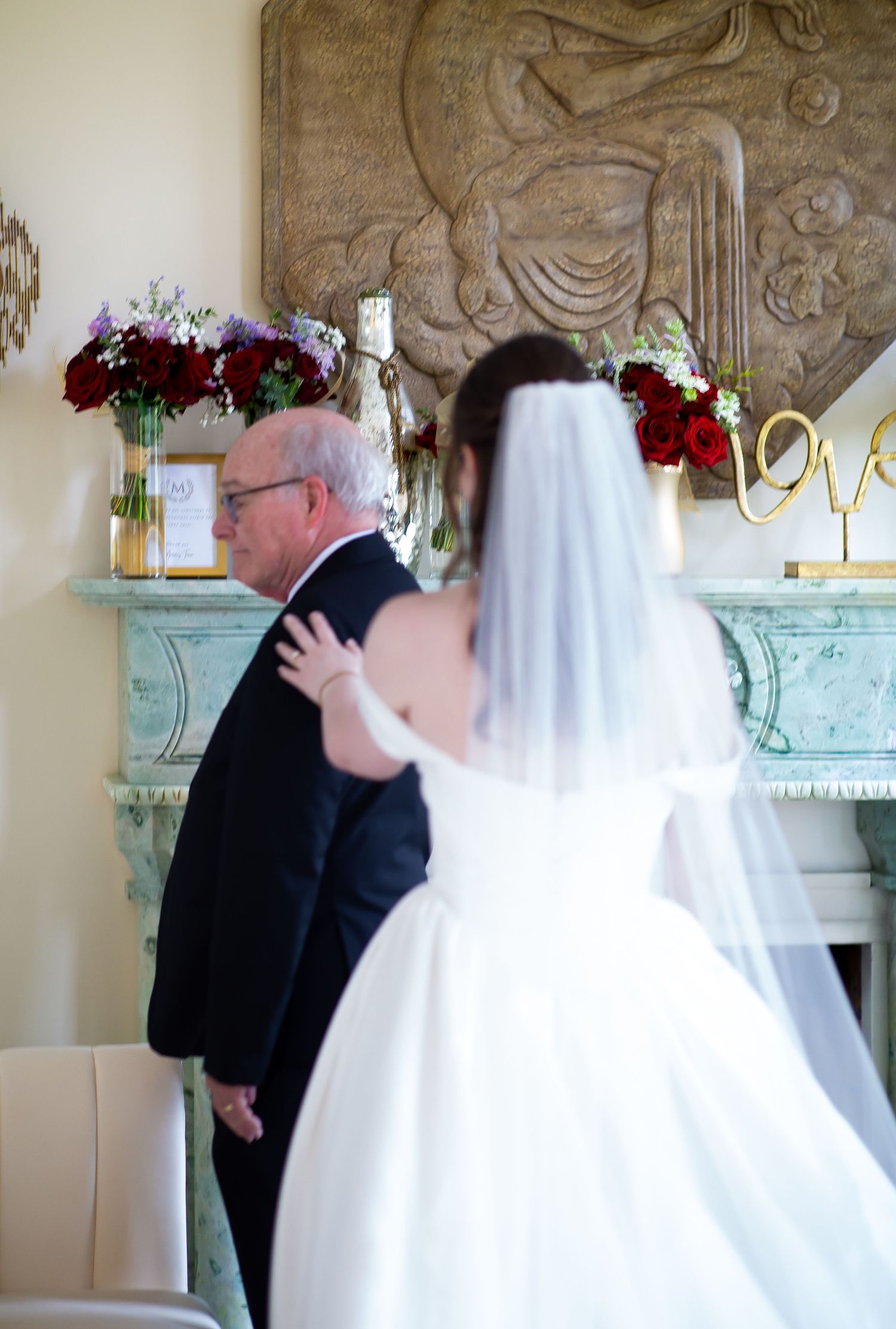 Bride in white ball gown and veil approaching her father near ornate fireplace.