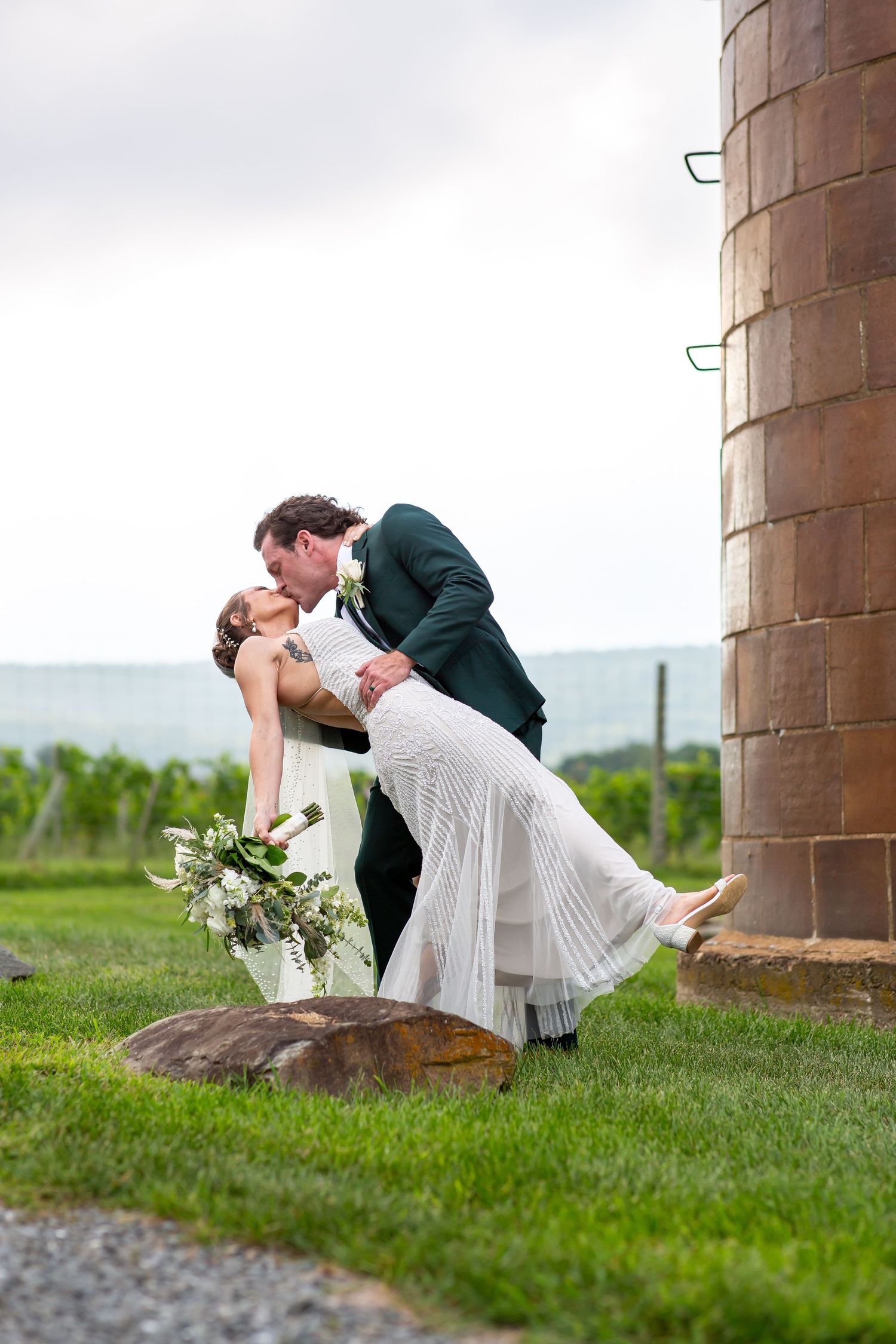 Bride and groom share a romantic dip kiss beside a rustic silo at a vineyard wedding venue.