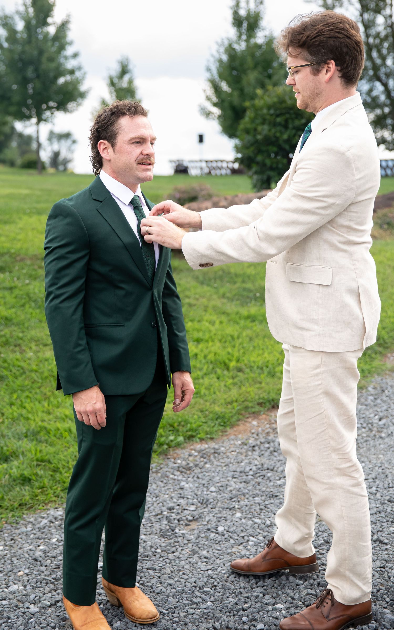 Two men in formal suits adjusting ties outdoors at a wedding venue with green grass background.