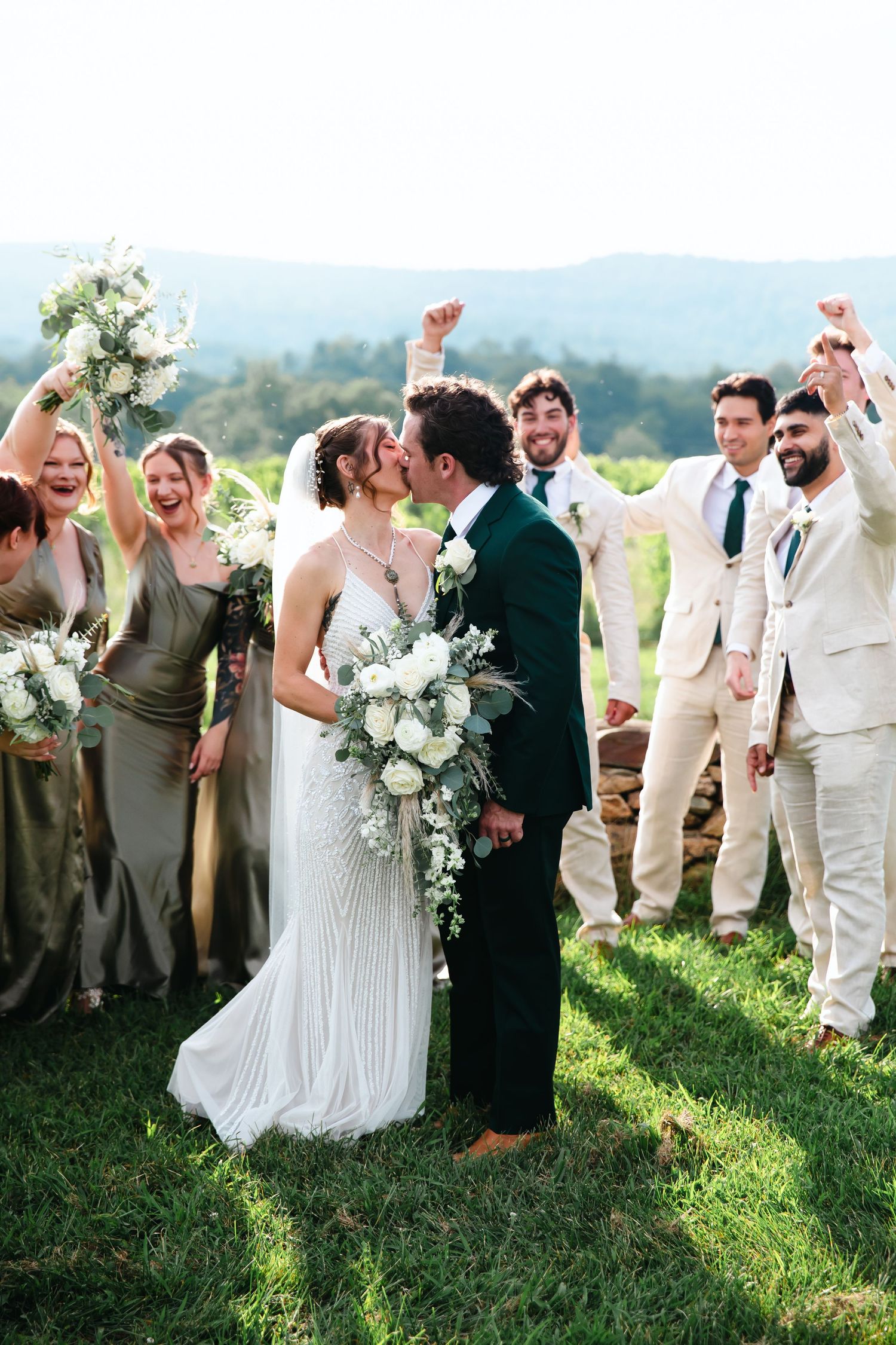 A bride and groom share a kiss surrounded by cheering wedding party members in sage green and ivory attire with mountain views.