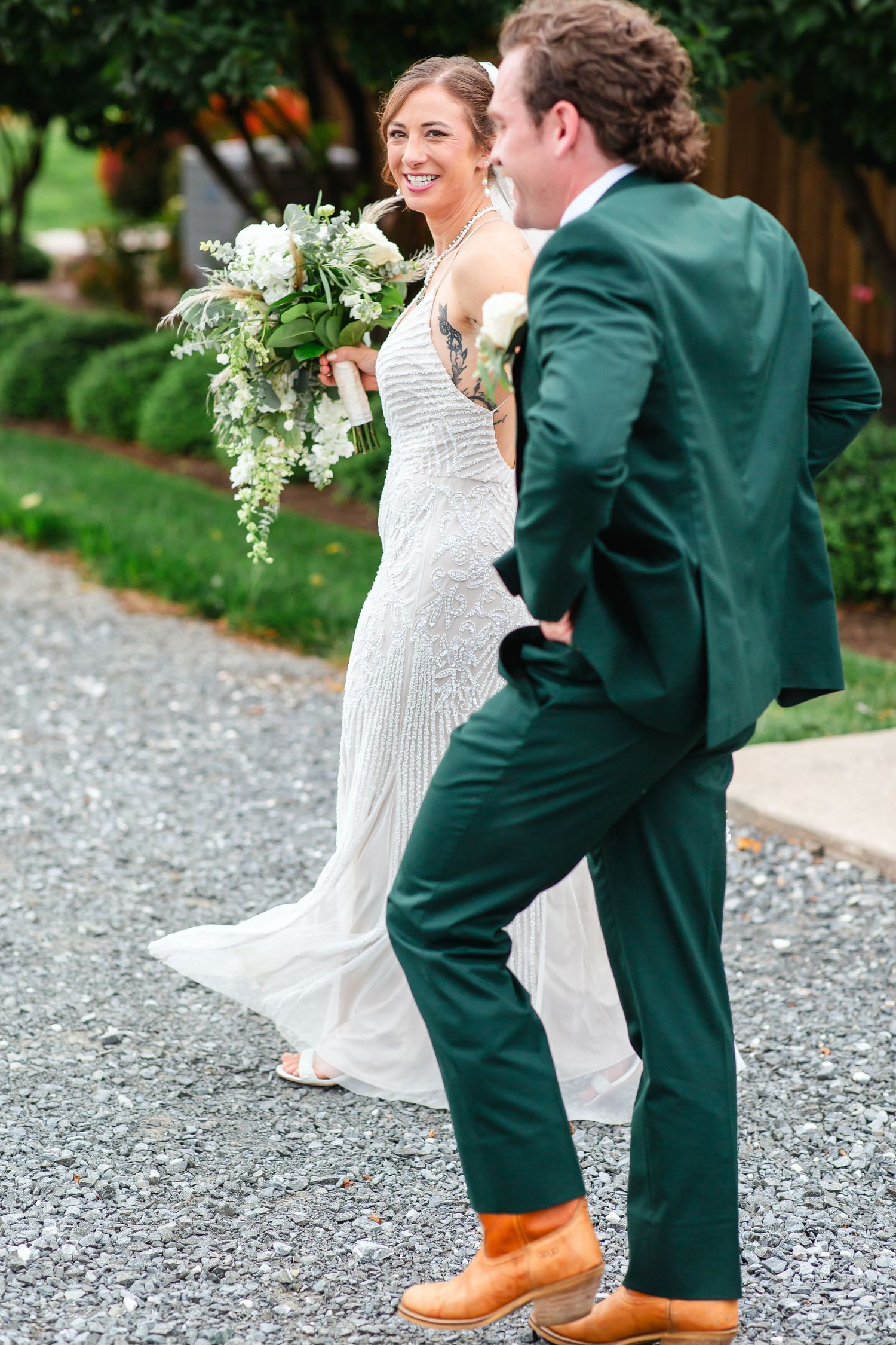Smiling bride in beaded gown holds white bouquet beside groom in green suit and brown boots on gravel path.
