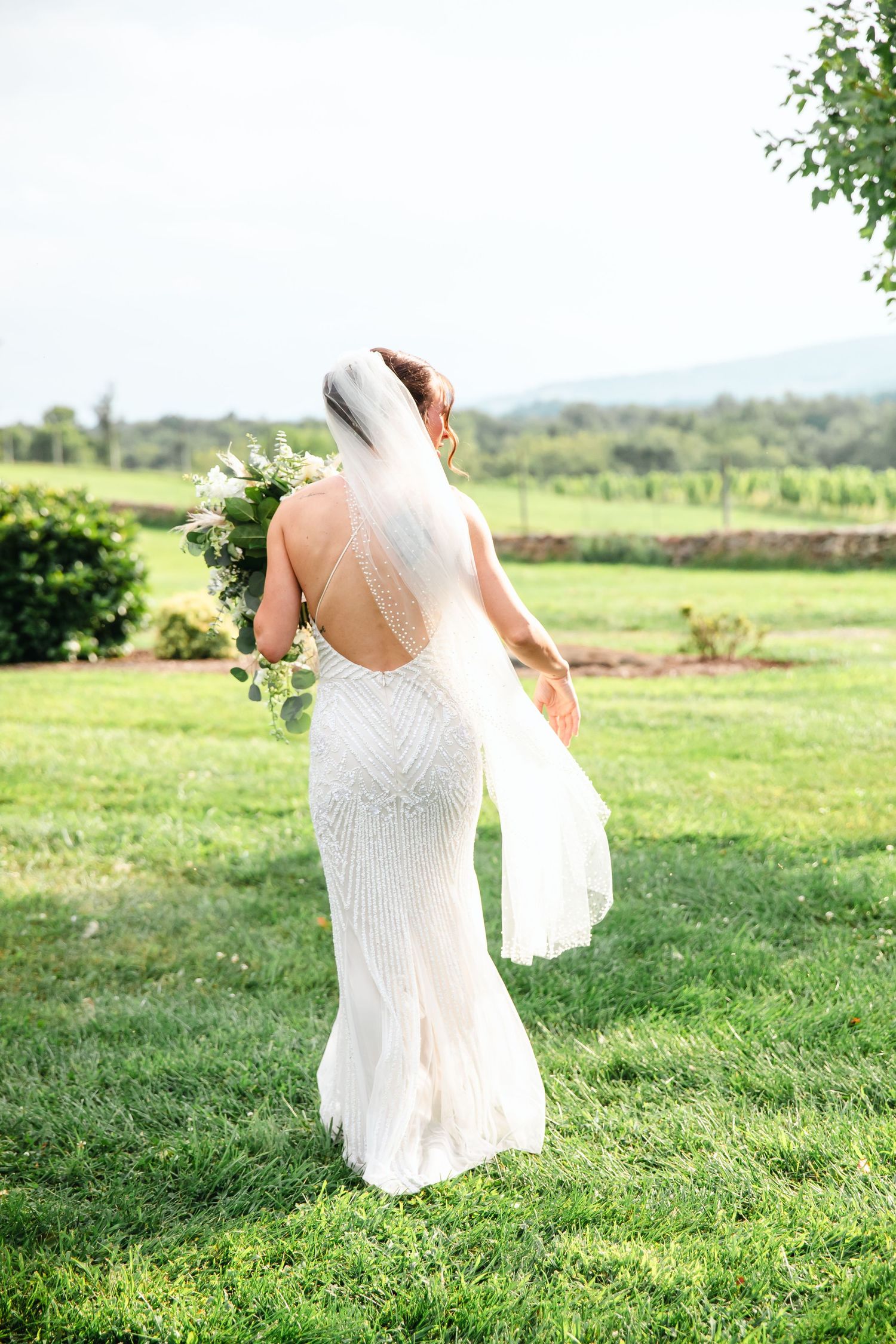 Bride in backless beaded gown and veil holds bouquet in lush green vineyard landscape.