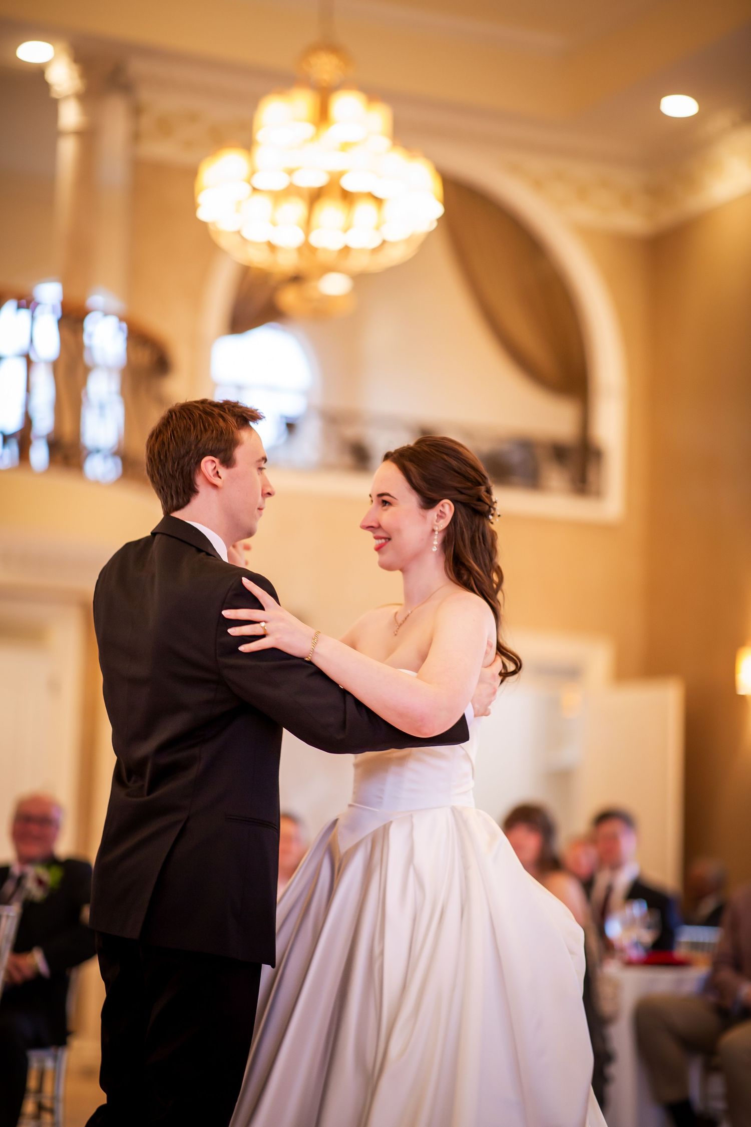 Smiling bride and groom share elegant first dance beneath a glowing chandelier at wedding reception.