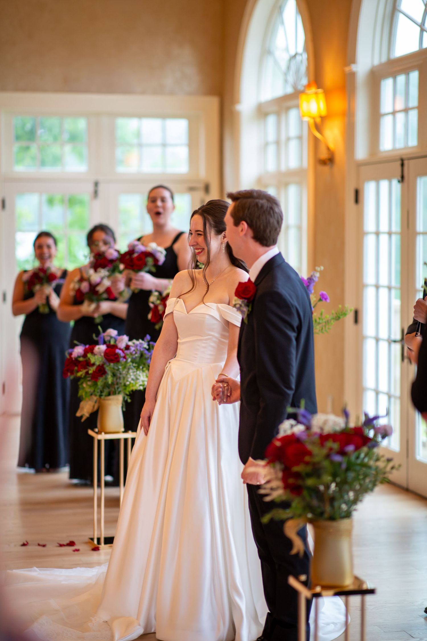 A joyful bride and groom share their first kiss at the altar surrounded by bridesmaids in black.