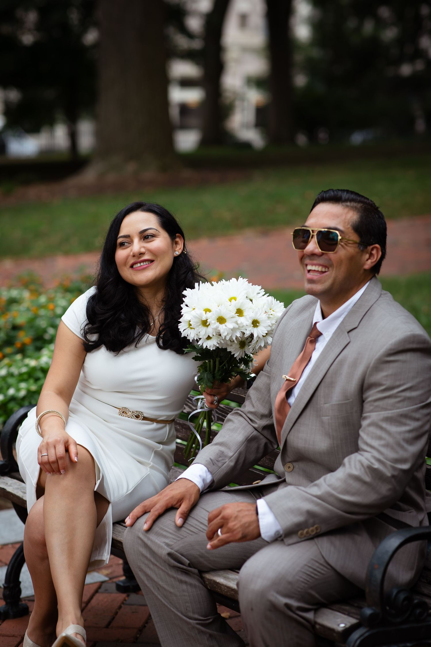 Smiling couple sitting on a park bench, woman in white dress holding white daisies, man in tan suit and sunglasses.