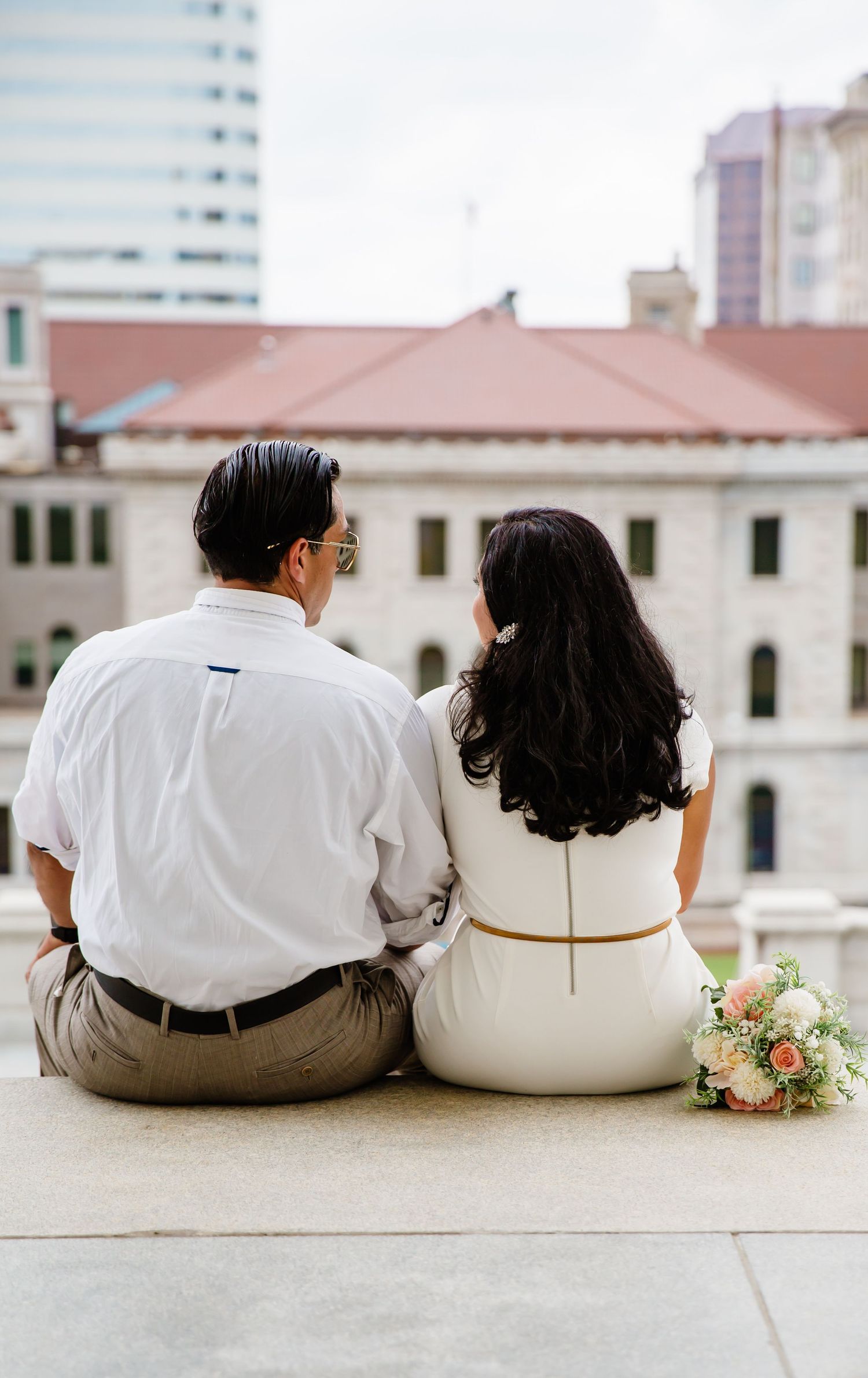 A couple in wedding attire sits together on a ledge, viewed from behind, with a bridal bouquet beside them.