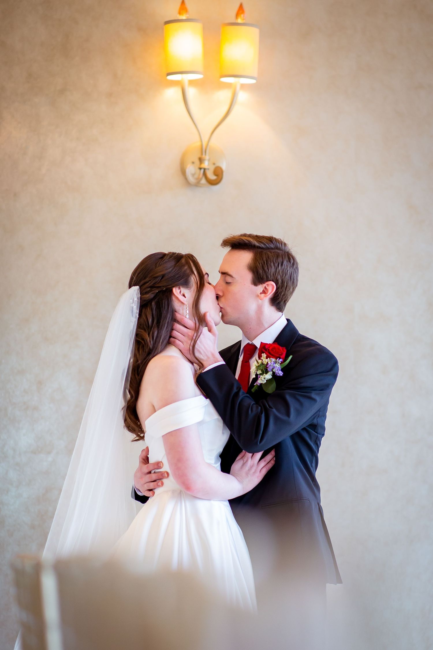 Bride and groom sharing first kiss in white off-shoulder gown under wall sconce light indoors.