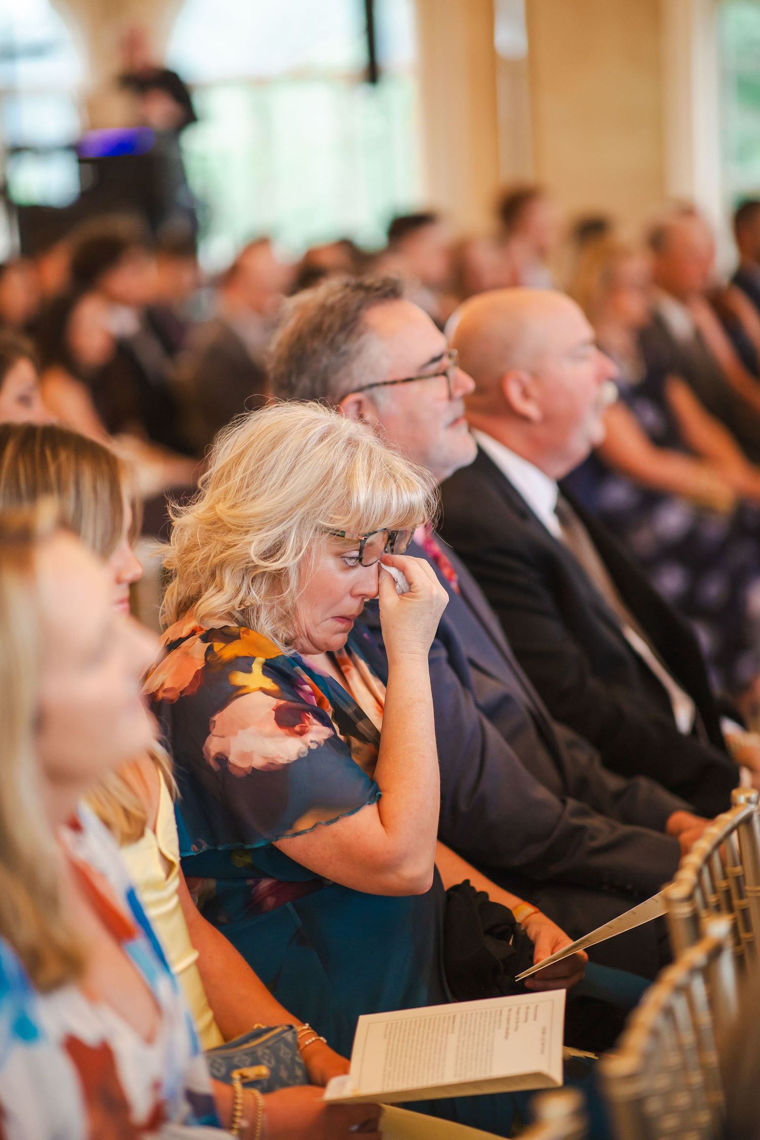 Emotional wedding guest wipes tears during heartfelt ceremony indoors.