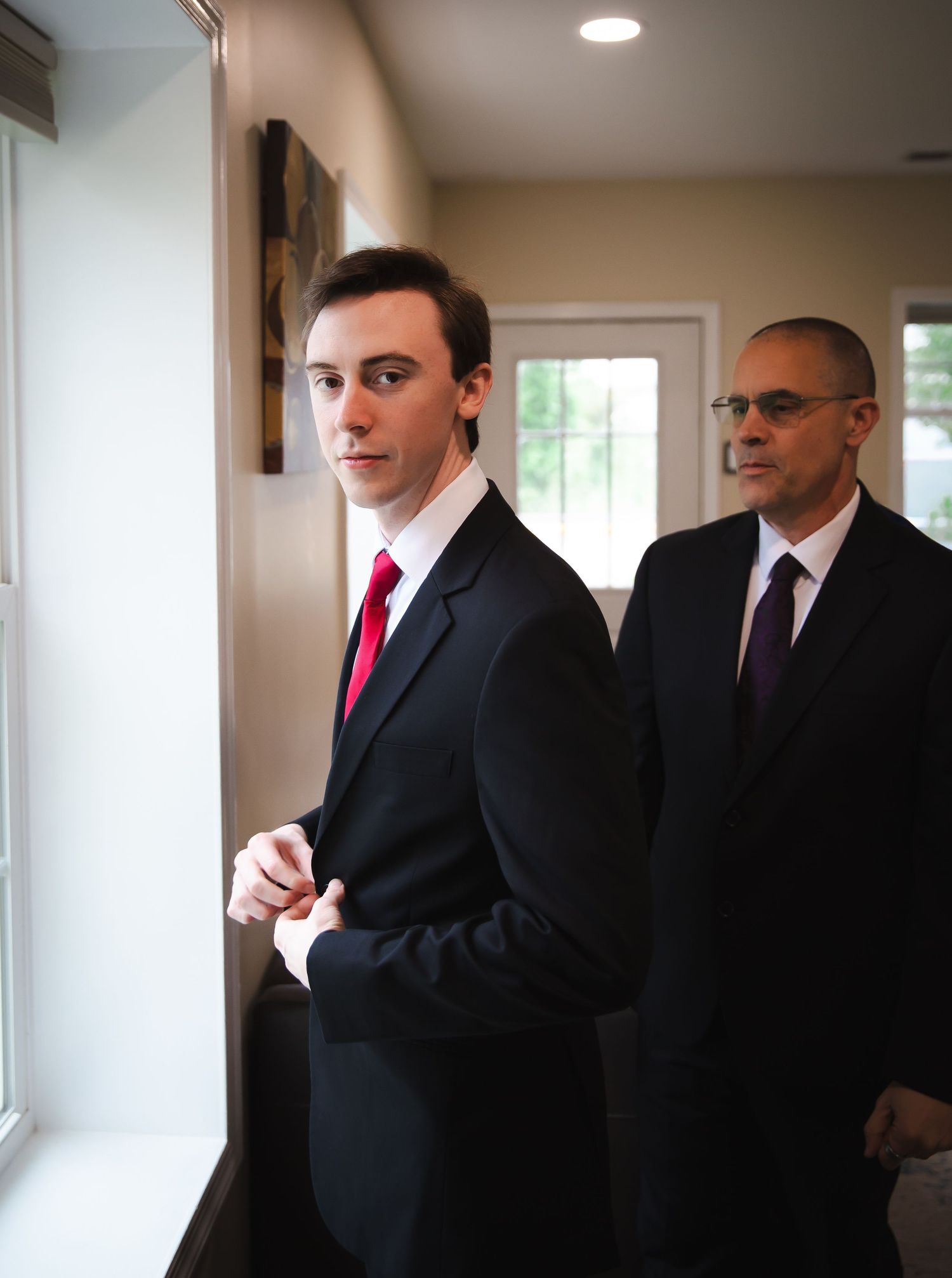 Young man in black suit with red tie standing by window with his father behind him.