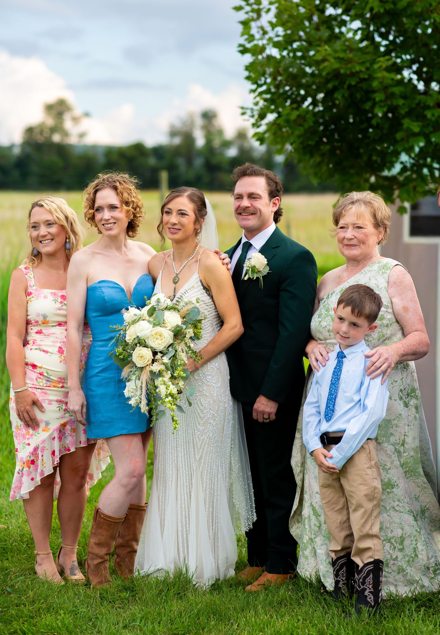 A bride and groom pose with family members outdoors at a wedding, with green fields in the background.