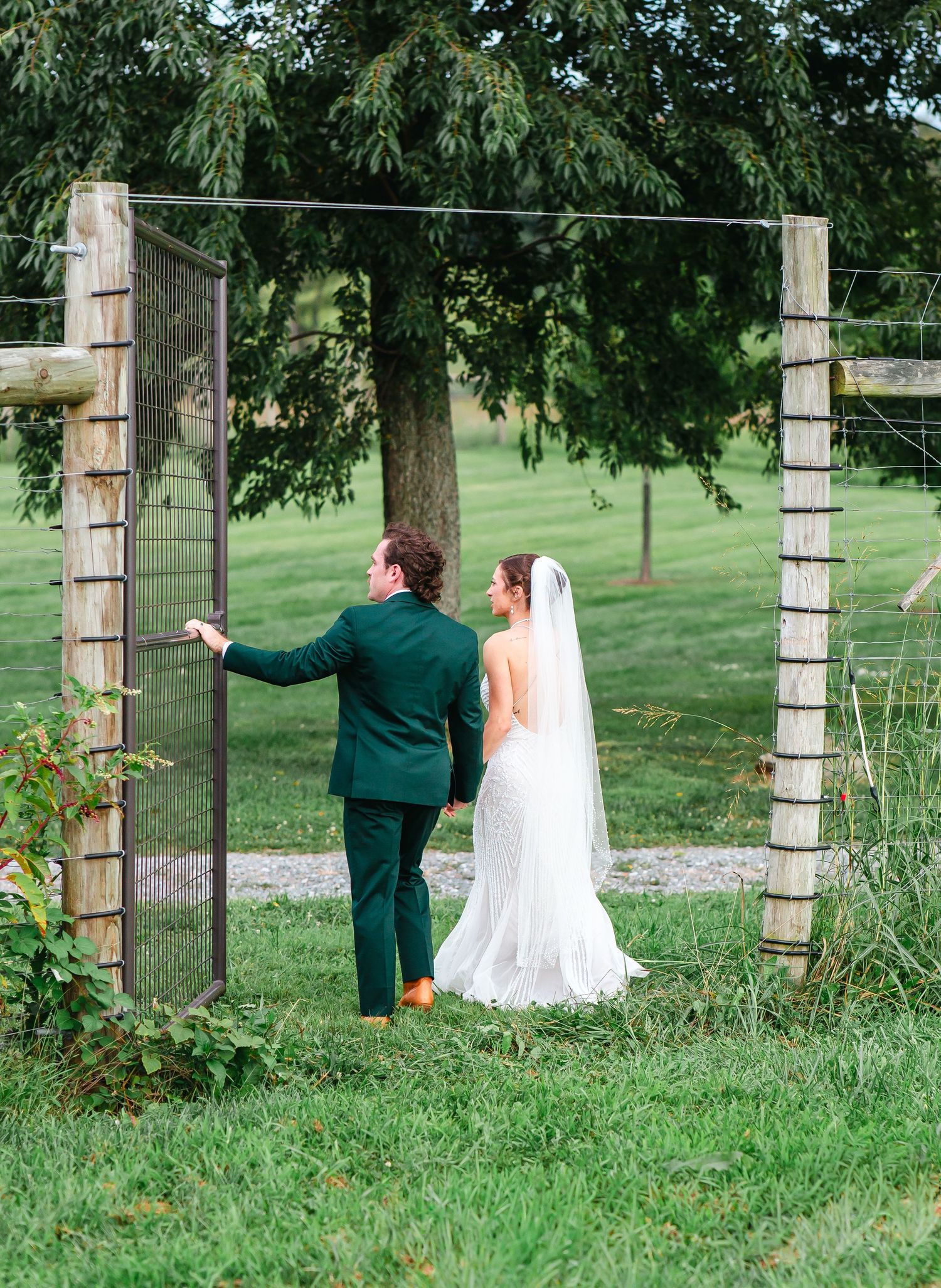 Bride in white gown and veil with groom in green suit walking through rustic gate on lush farm wedding venue.