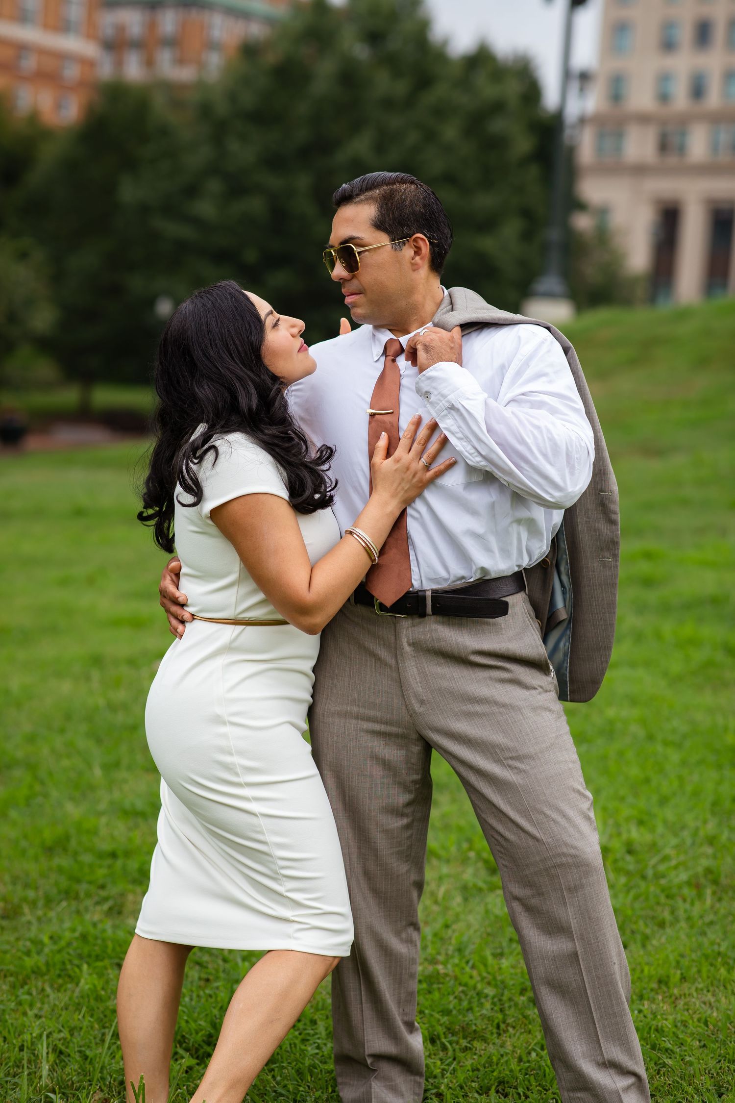 Couple in formal attire embracing outdoors, woman in white dress, man in gray suit with pink tie and sunglasses.