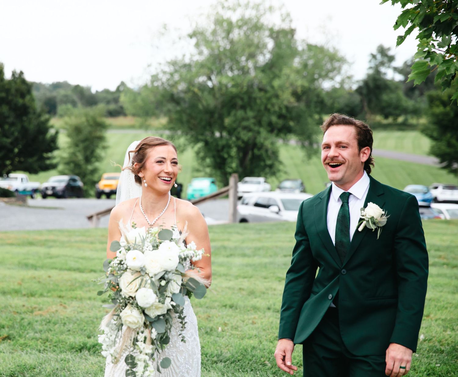 A smiling bride holding a white bouquet and groom in a green suit walk together on a green lawn at their outdoor wedding.