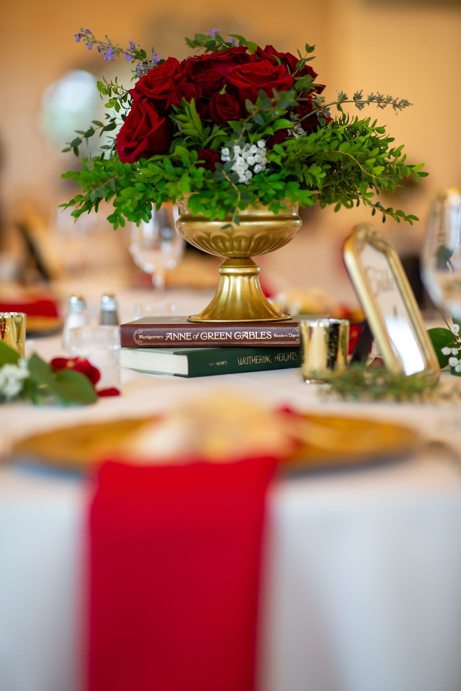 Gold vase centerpiece with red roses and greenery atop classic novels on white wedding table.