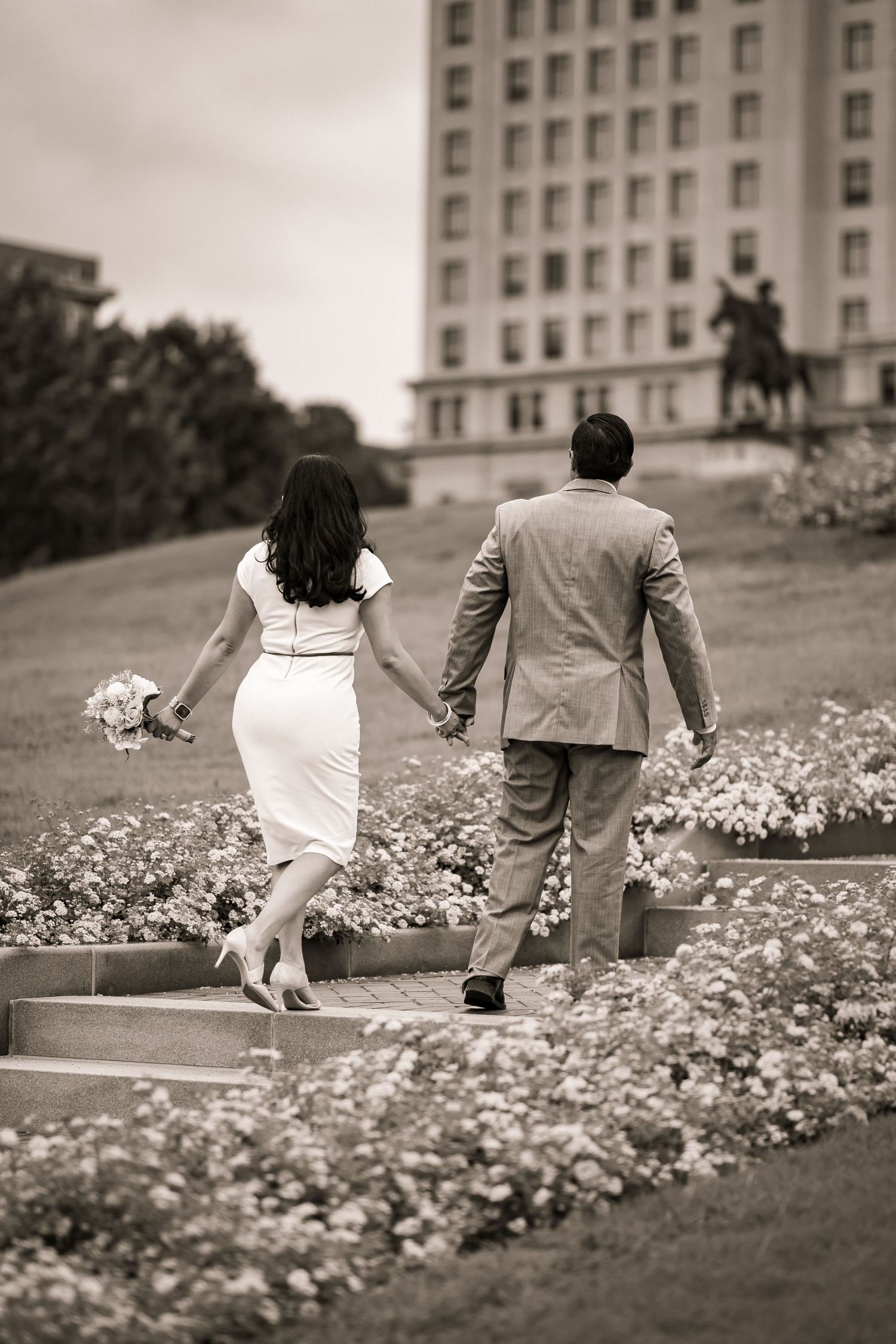 Black and white photo of a bride and groom holding hands walking up steps through a flower garden.