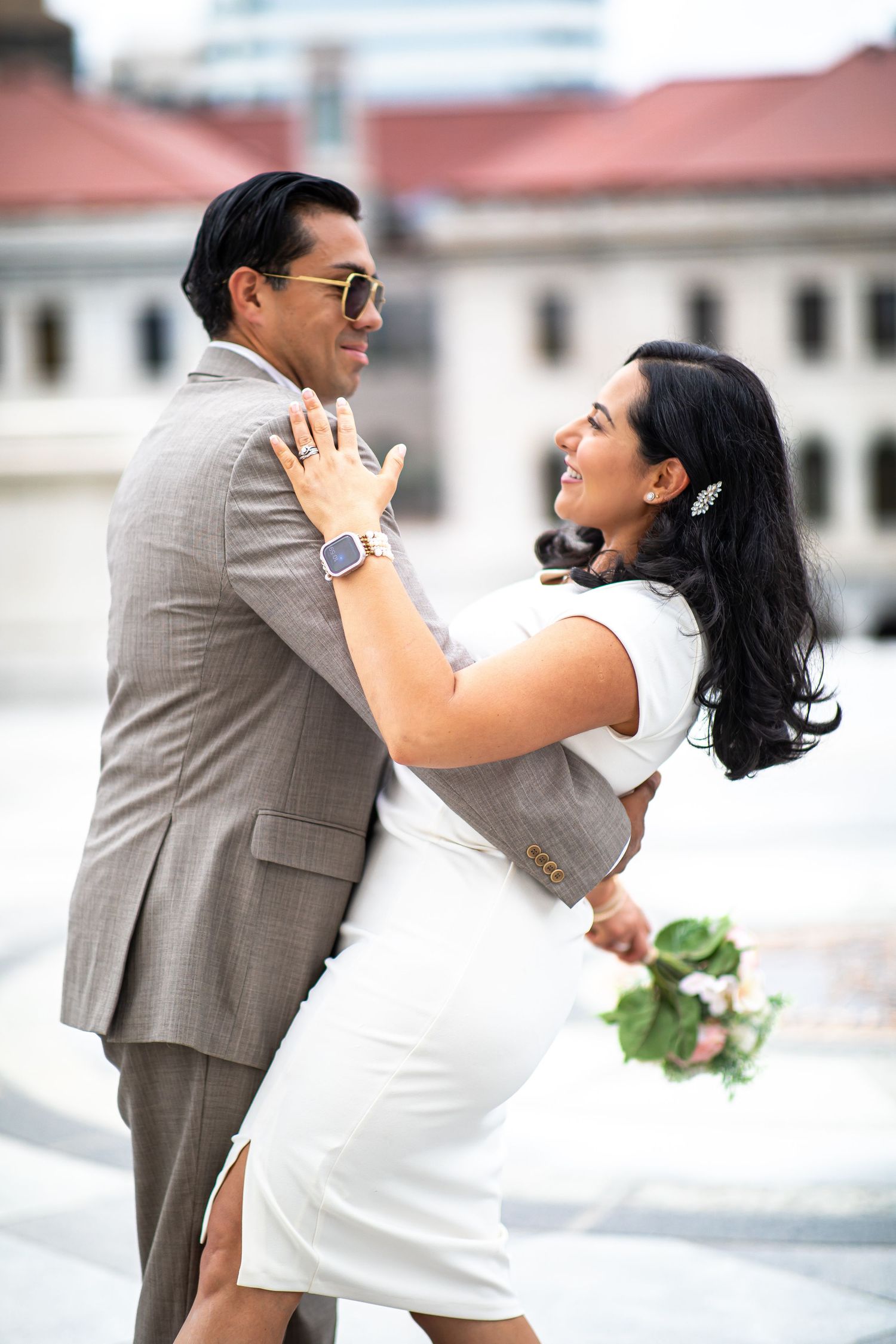 A bride in a white dress holds flowers while embracing a groom in a gray suit with sunglasses outdoors.