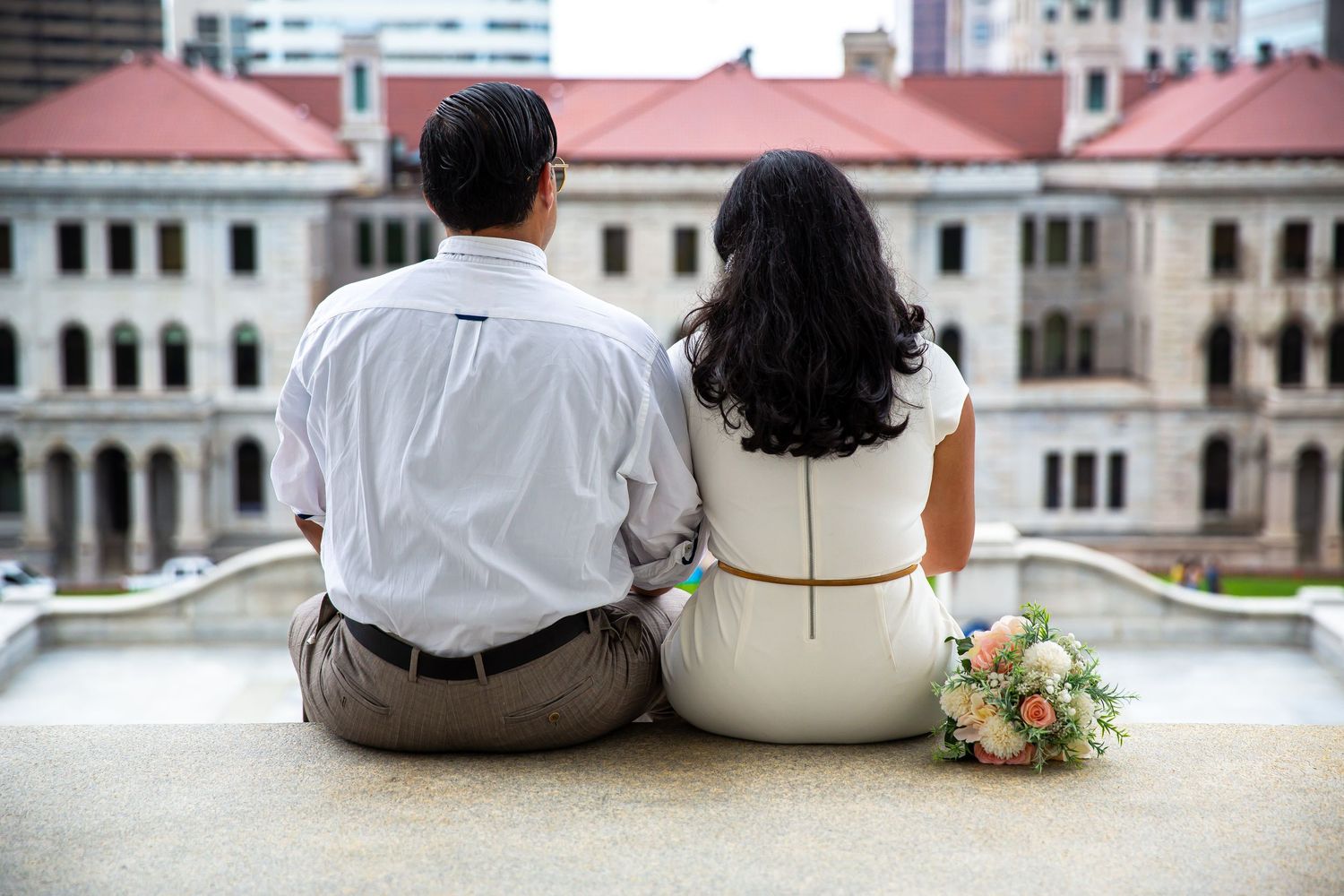Couple sitting together on stone ledge with bridal bouquet, backs to camera, facing historic building.