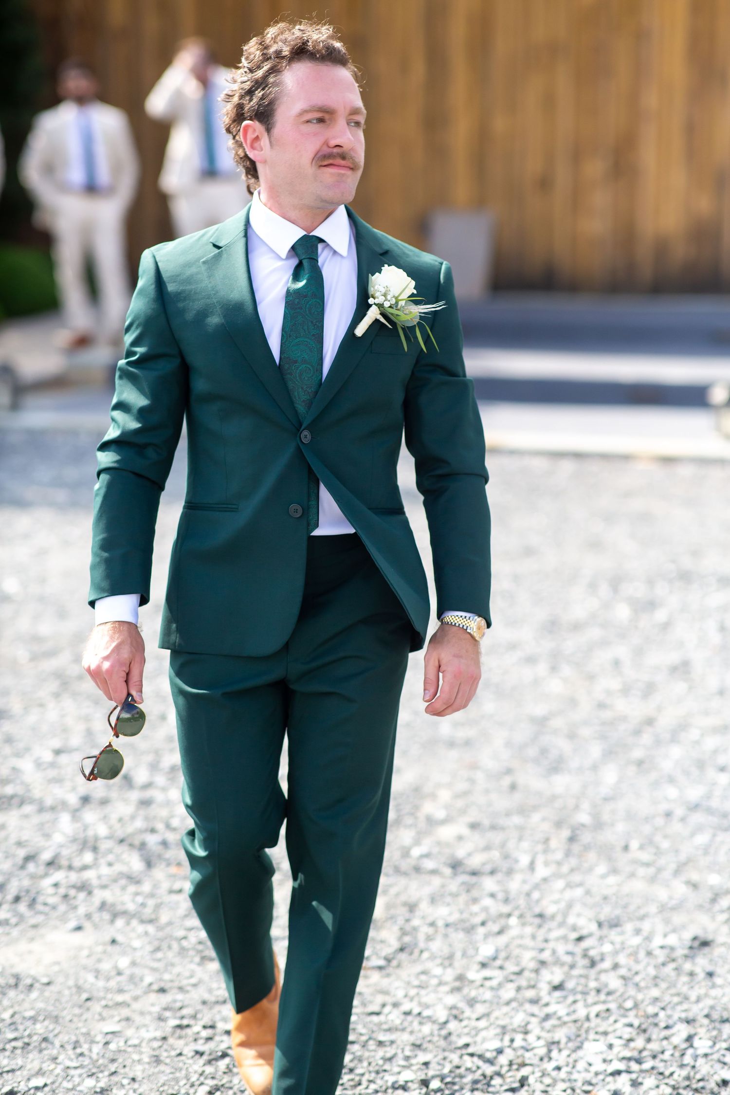 Groom in dark green suit with boutonniere walks on gravel path at outdoor wedding venue.