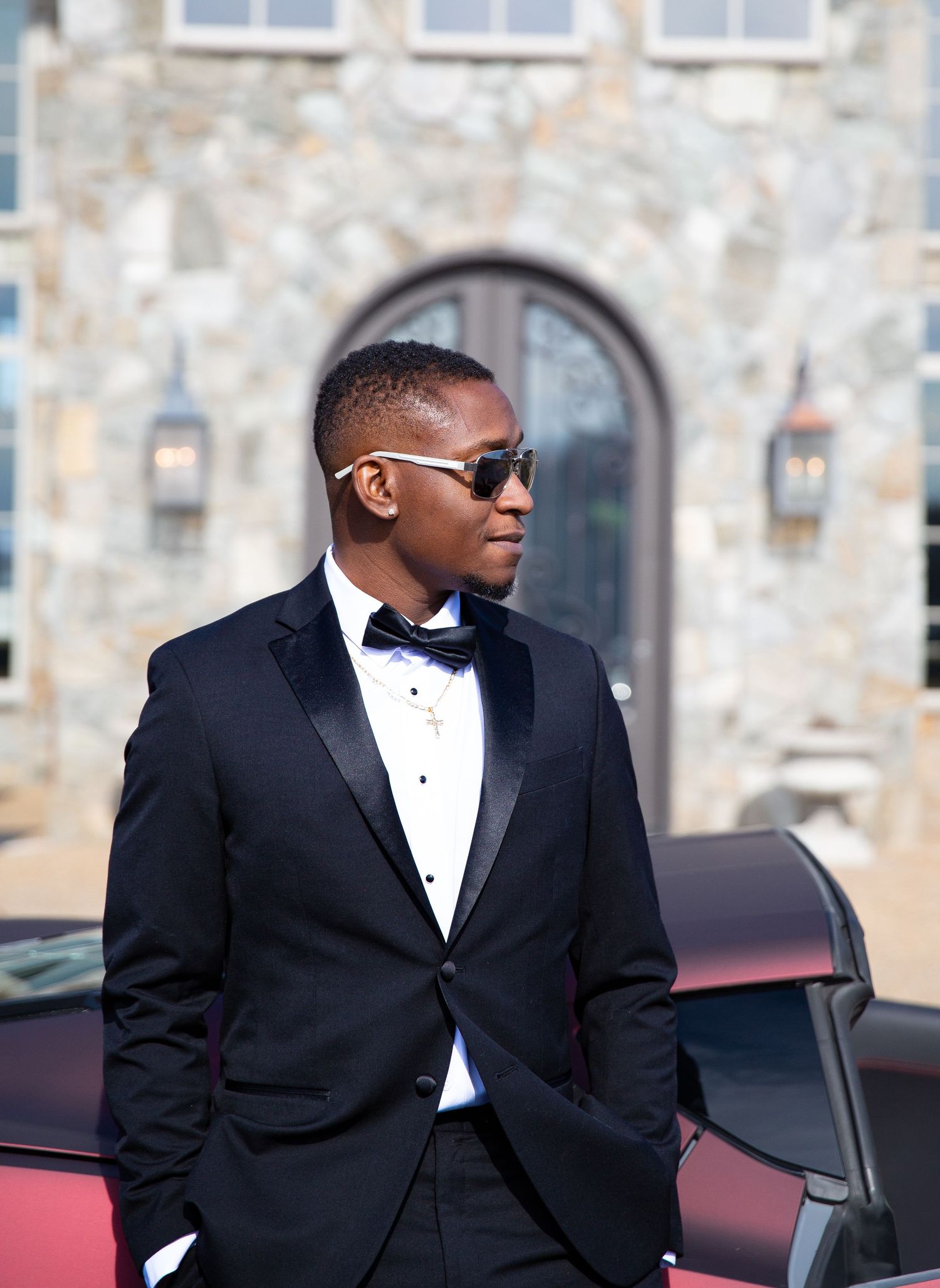 Young Black man in black tuxedo with sunglasses leaning on car outside stone building.