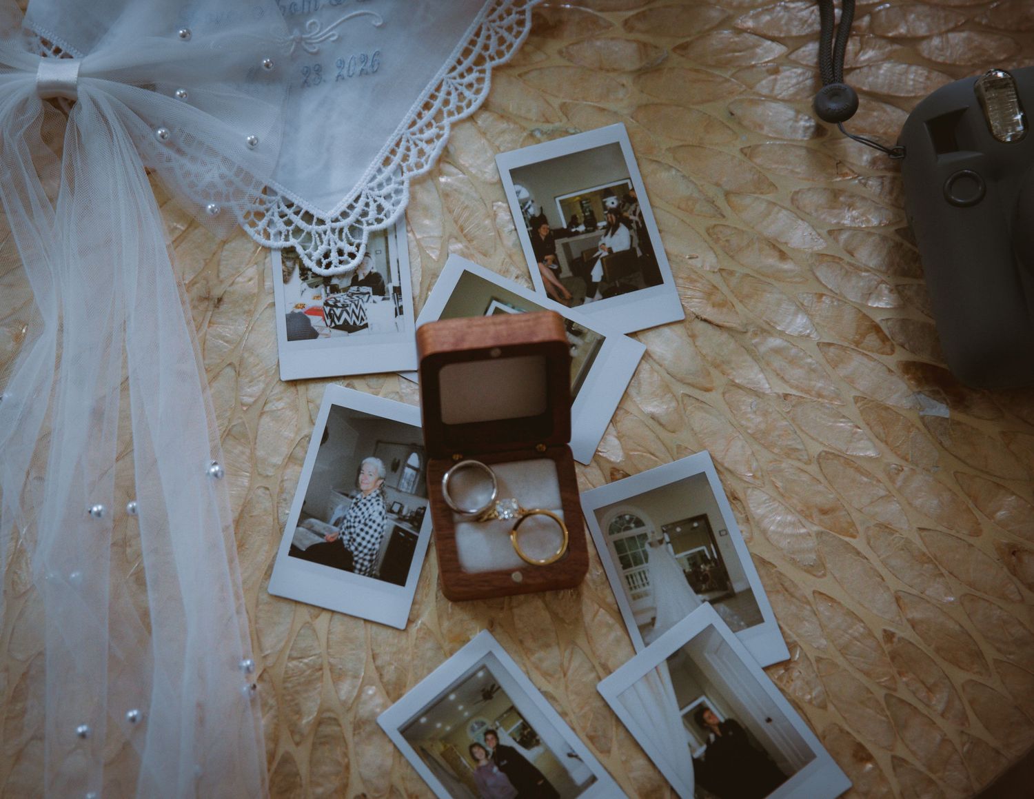 Wedding rings in wooden box surrounded by Polaroid photos and pearl-edged bridal veil on patterned floor.