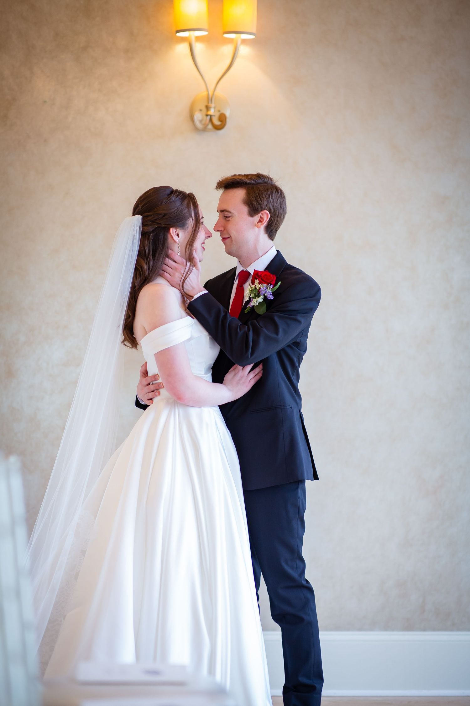 Bride and groom share tender moment embracing indoors, bride wearing white off-shoulder gown and veil.