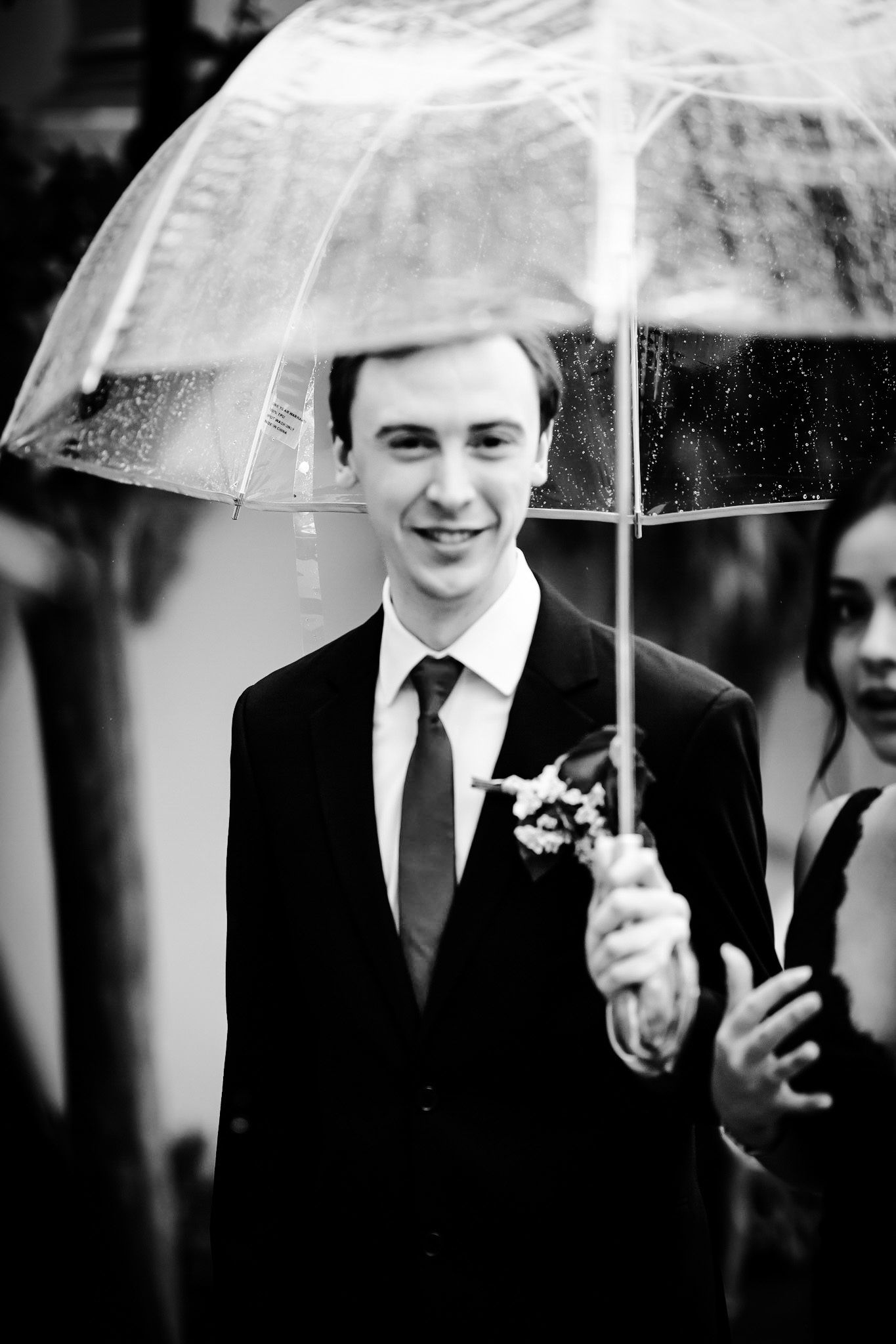 Smiling groom in black suit with boutonniere holding clear umbrella in black and white wedding photo.