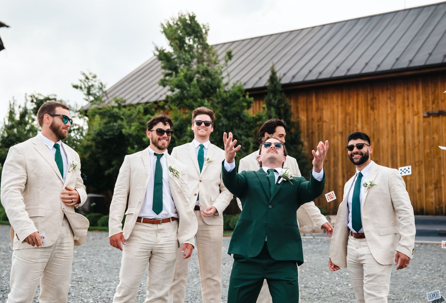 Groom in dark green suit with groomsmen in beige suits and teal ties celebrating outdoors at a rustic wedding venue.
