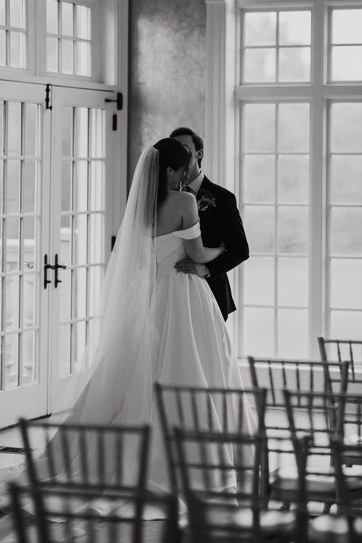 Bride and groom kissing in black and white photo near French doors with wedding chairs in foreground.