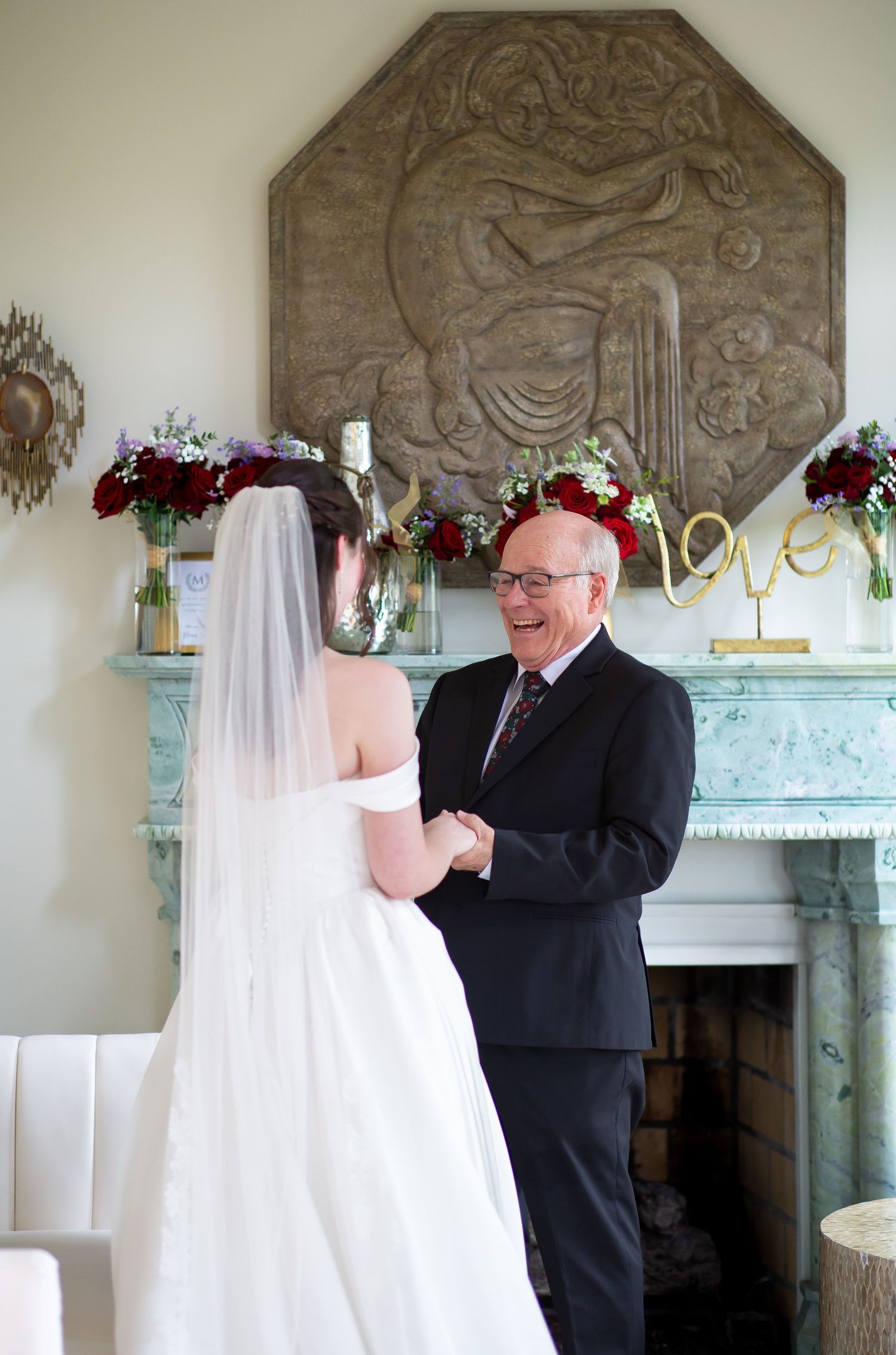 Bride in off-shoulder gown holding hands with her smiling father near elegant fireplace.