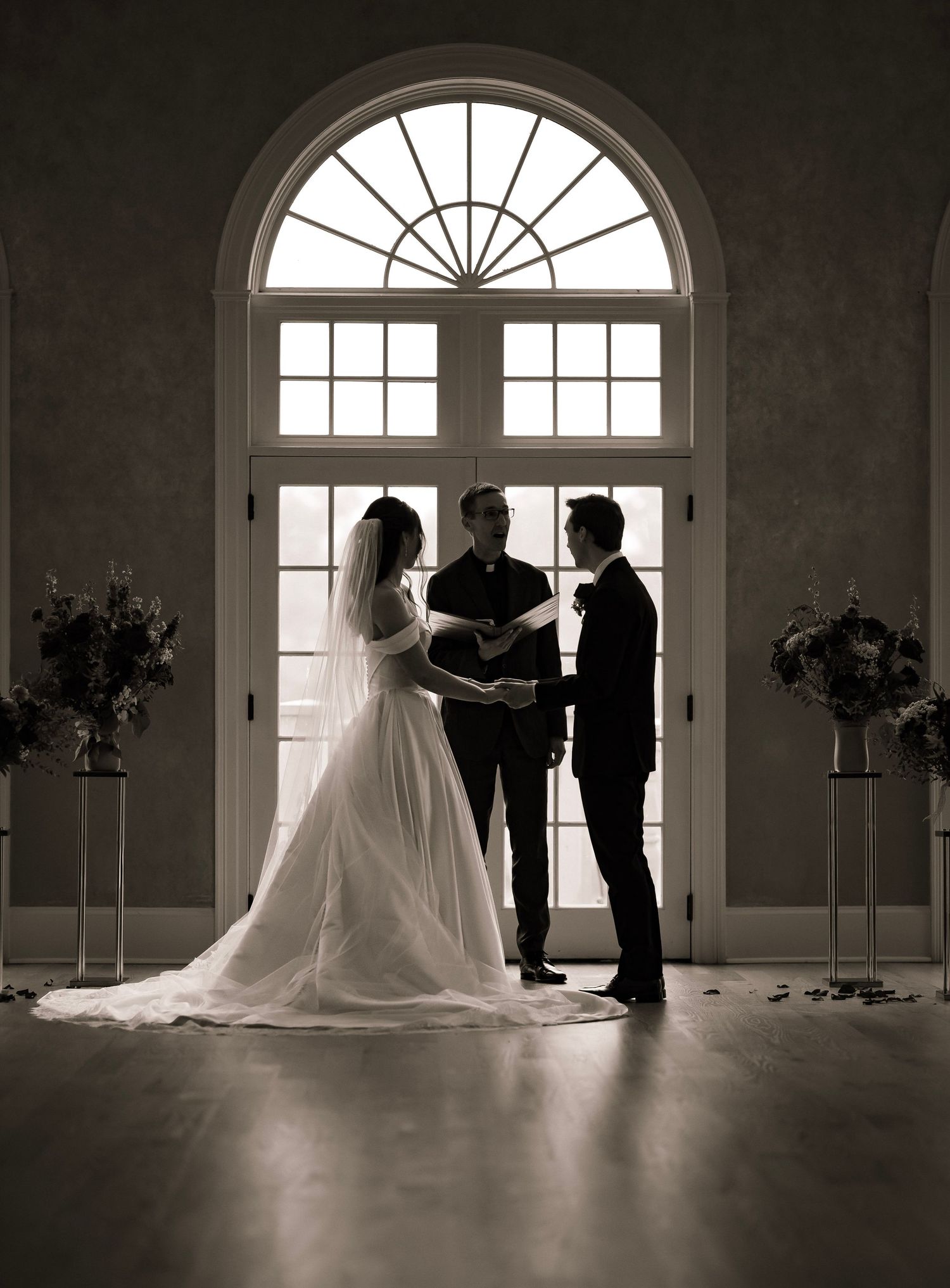Black and white photo of bride and groom exchanging vows before officiant in elegant arched window venue.