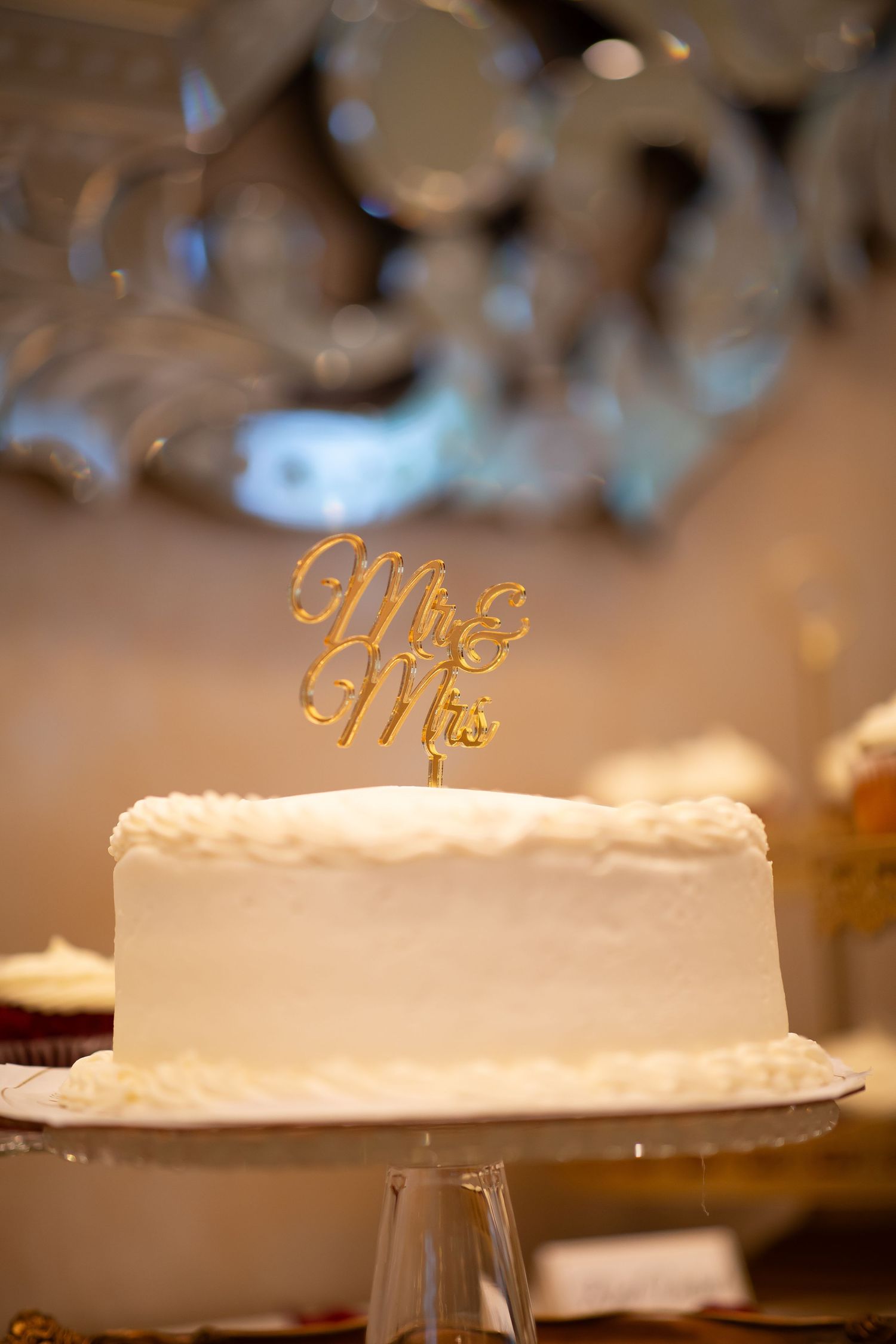 White wedding cake with gold Mr and Mrs topper displayed on crystal cake stand at reception.